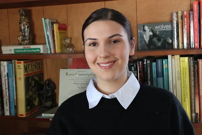 A young woman with dark hair, wearing a white collared shirt and black sweater, standing in front of a bookshelf filled with books and decorative items.