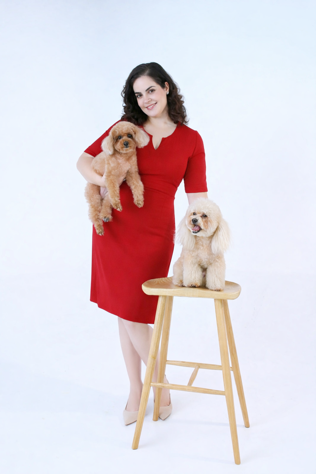A woman in a red dress holding a small poodle and standing next to a chair with a light-colored poodle on top, all set against a plain white background.
