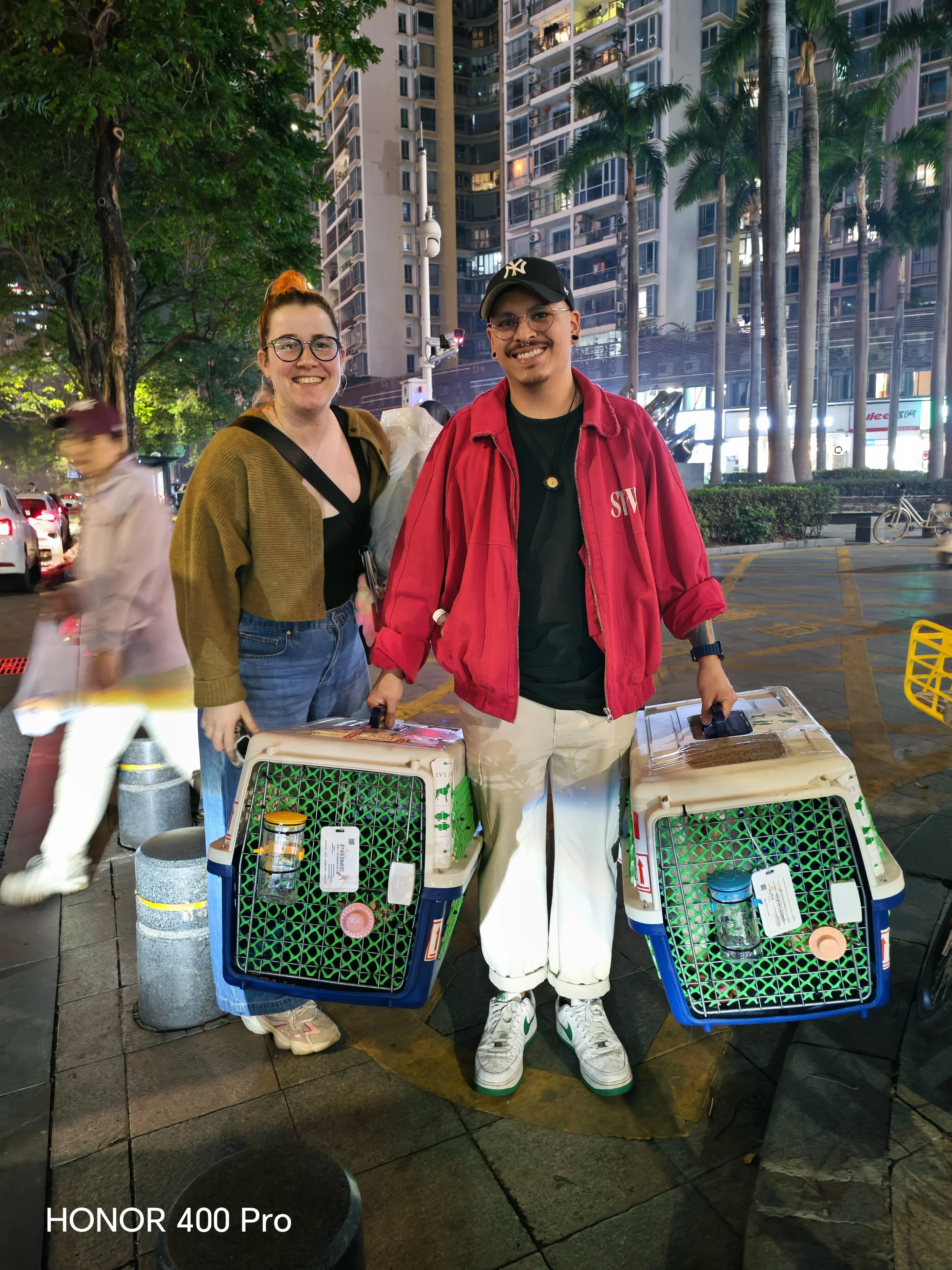 Two people standing outdoors at night holding pet carriers, with city buildings and palm trees in the background.