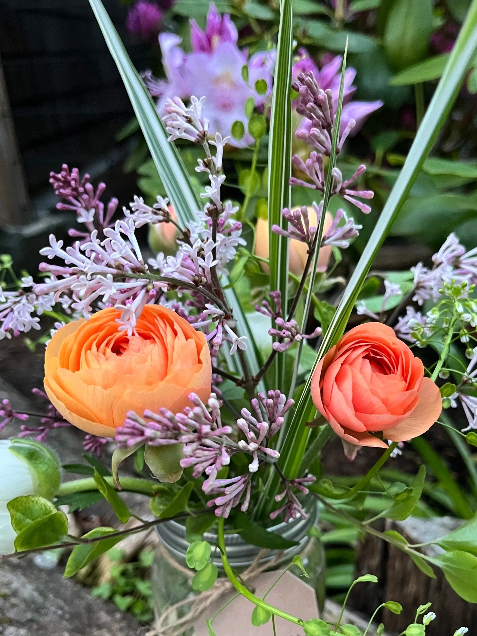 A small bouquet of fresh flowers in a glass jar, featuring orange ranunculus, purple and white lilac-like flowers, and other green foliage, with a handwritten tag attached.