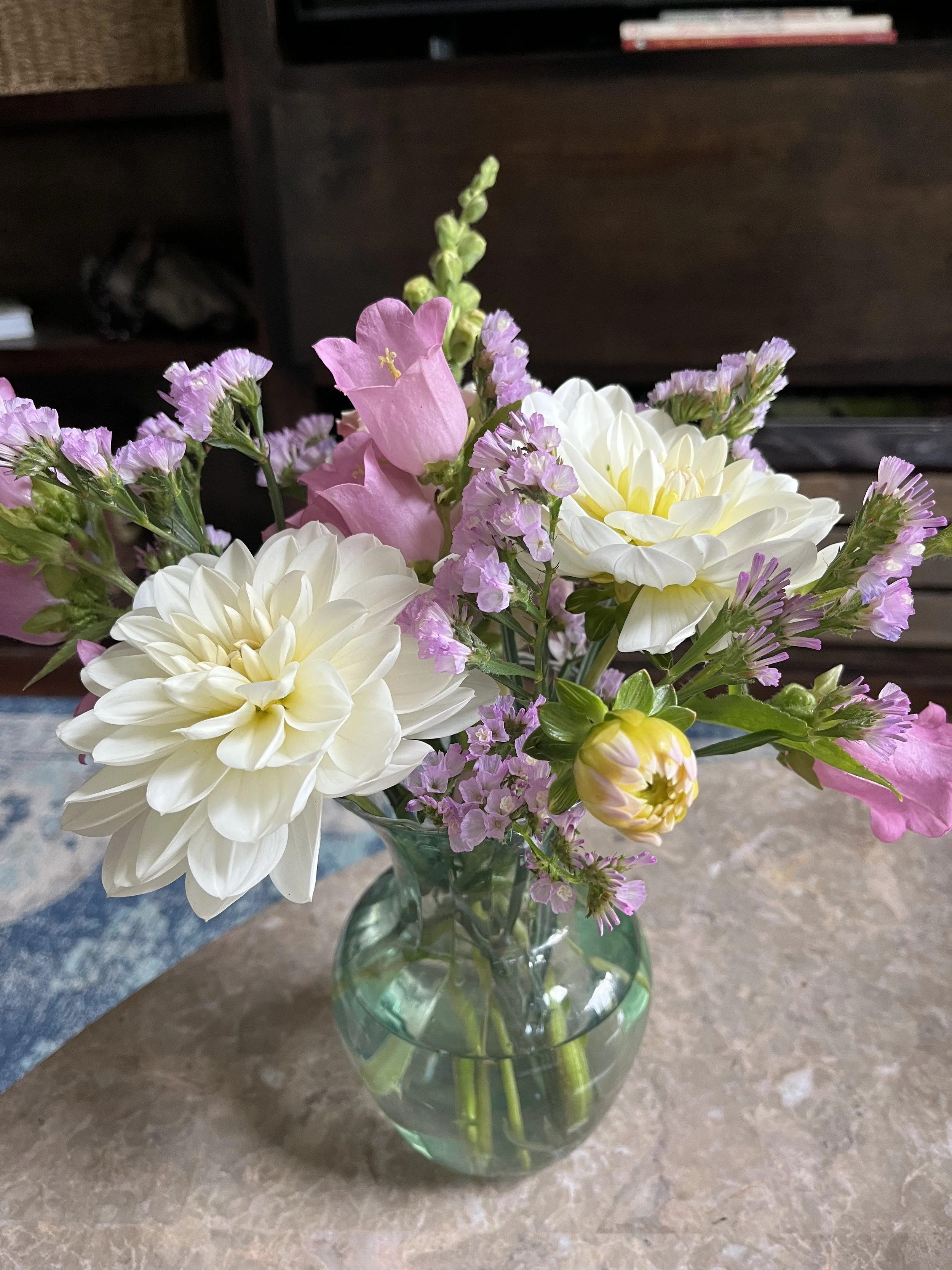 Bouquet of colorful flowers including pink dahlias, white petunias, purple statice, and other blooms in a glass vase with a tag that reads "Blumenhotte" on a wooden surface