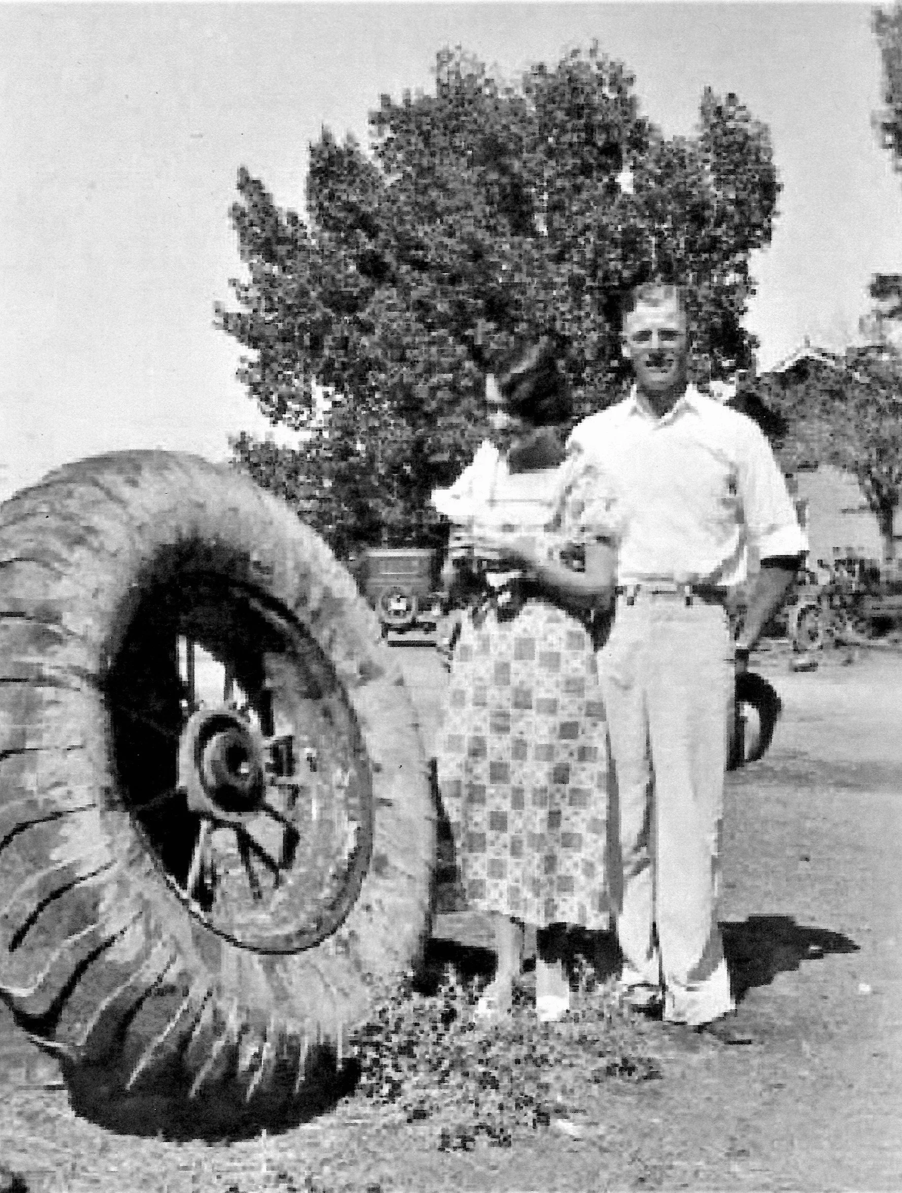 Black and white photo of a woman standing next to a large, vintage tractor in an outdoor area with trees in the background. She is wearing sunglasses, a light-colored shirt, and a skirt.