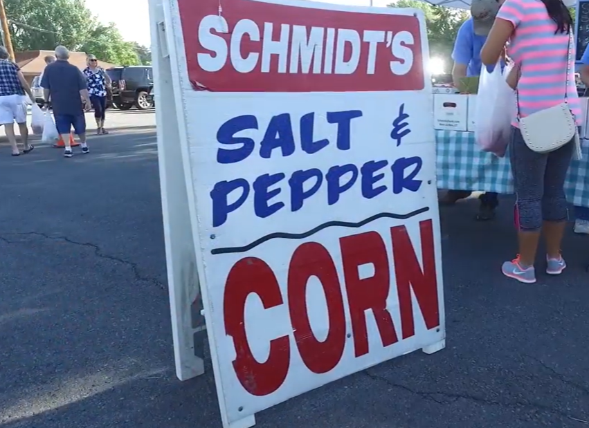 A white signboard advertising Schmidt's salt and pepper corn at an outdoor market, with people shopping in the background and trees visible.