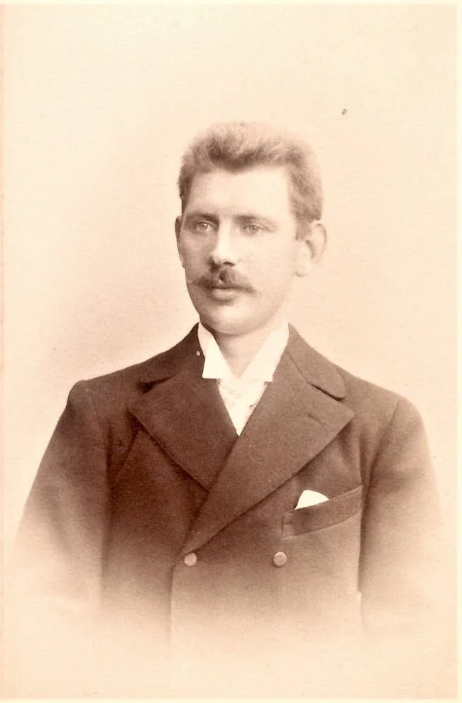 Black and white vintage portrait of John Schmidt taken in Germany with short hair and a mustache, wearing a suit with a v-neck shirt and a pocket square.