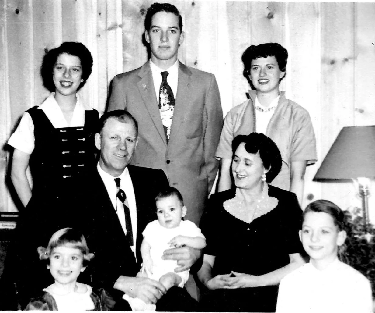 A black and white family portrait with nine people, including three children, two men, and four women, standing and sitting in a room with wooden panel walls. The group appears to be from the mid-20th century.
