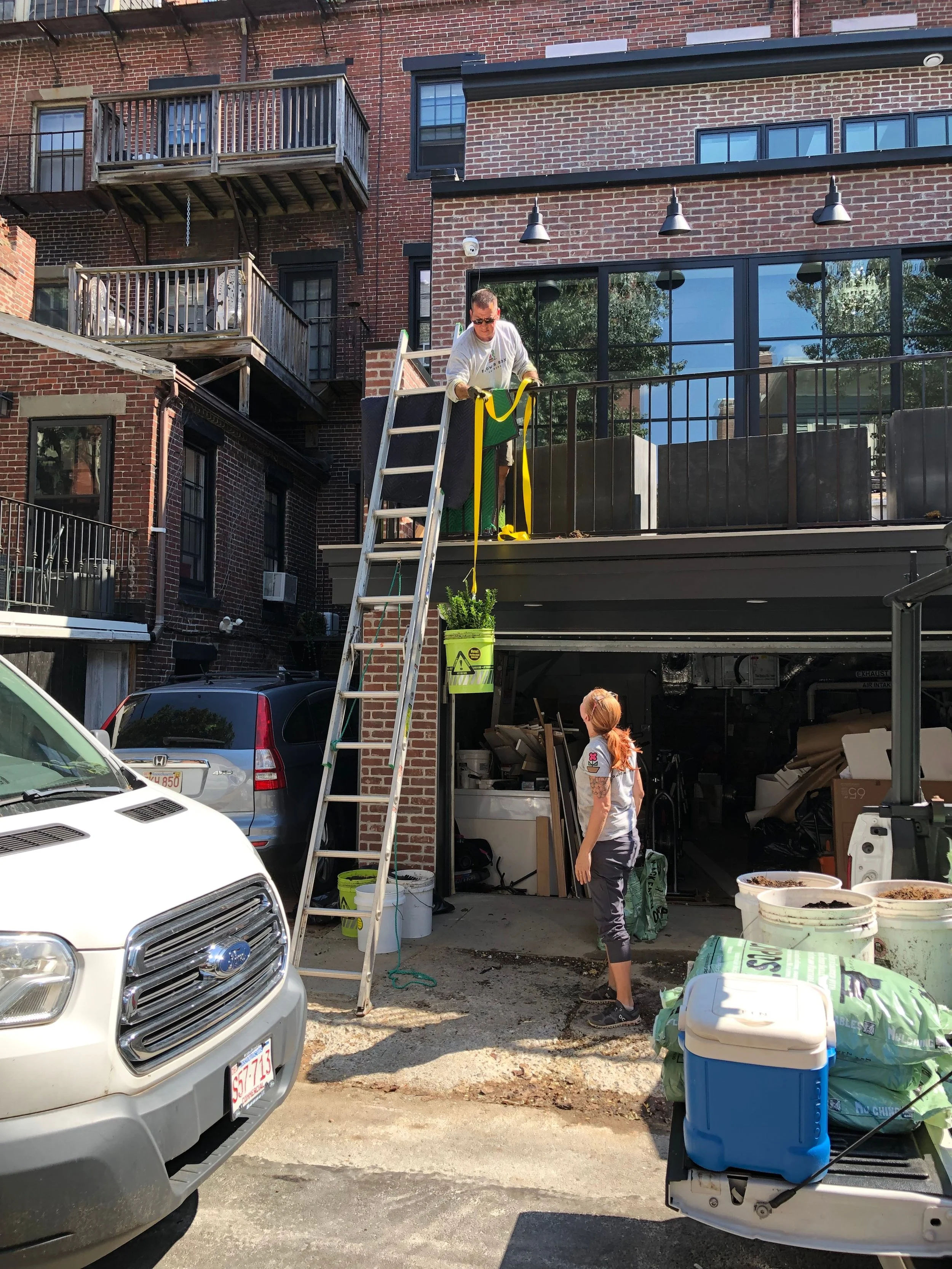 A person standing on a ladder near a building's balcony, holding a yellow and green object while another person watches from below in an outdoor setting.