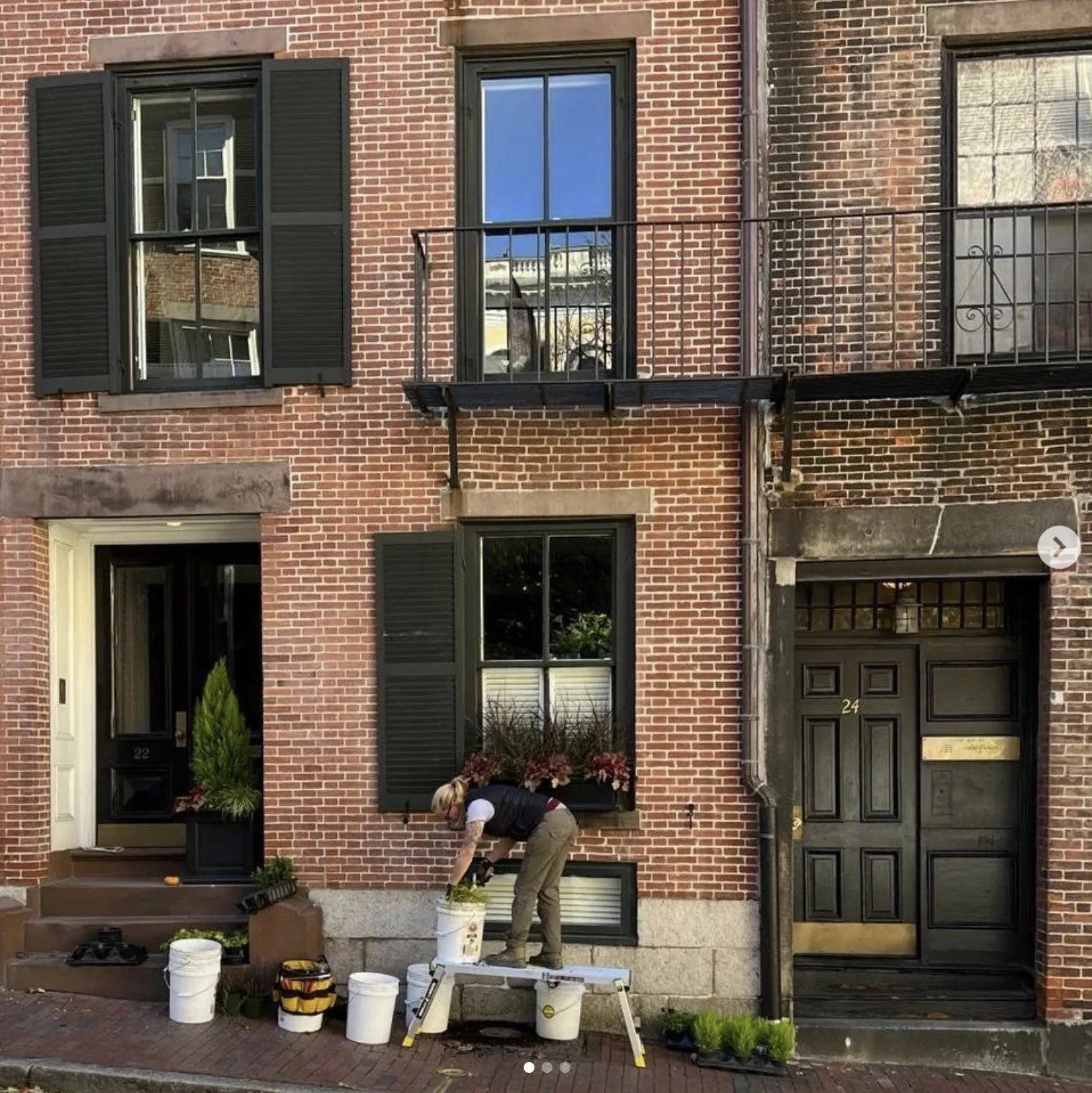 Person gardening outside a brick townhouse with black shutters and doors, using buckets and tools during the day.
