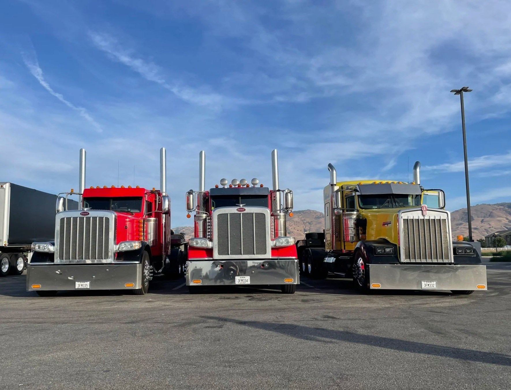 Three large semi-trucks parked side by side in a lot against a backdrop of mountains and a blue sky with clouds.
