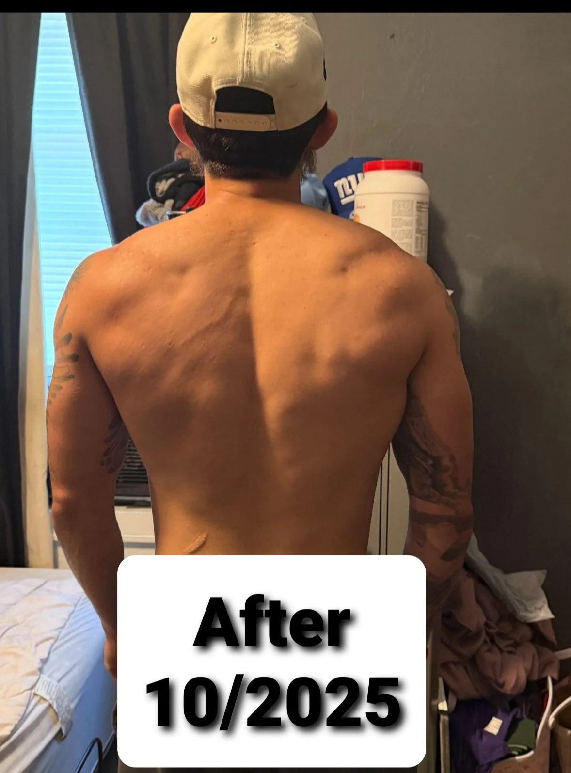 Back view of a shirtless man with tattoos, wearing a beige baseball cap, standing in a bedroom next to a gray wall with laundry and a large container of protein powder.