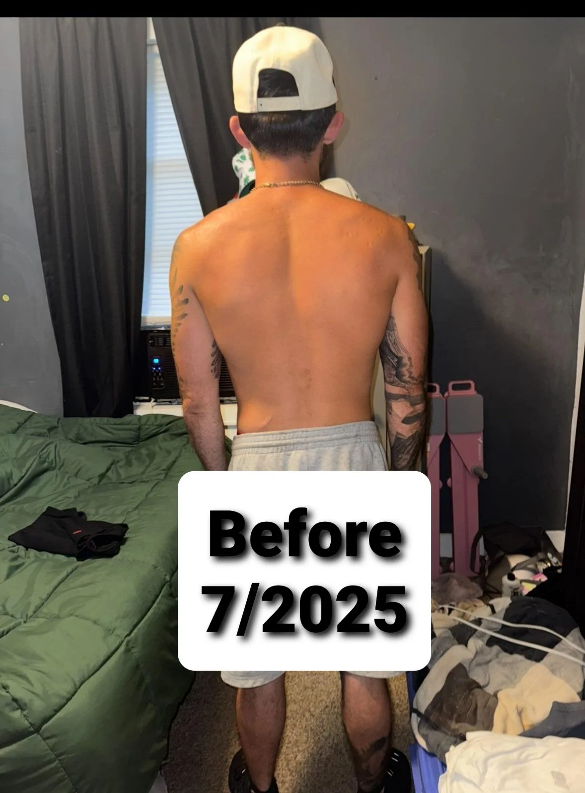 Back of a shirtless young man wearing a white baseball cap, standing in a bedroom with dark walls and black curtains. There are tattoos on both arms, and he is wearing grey shorts. A bed with a green comforter, some clothes, and miscellaneous items are visible in the room. The text 'Before 7/2025' is overlaid on the image.