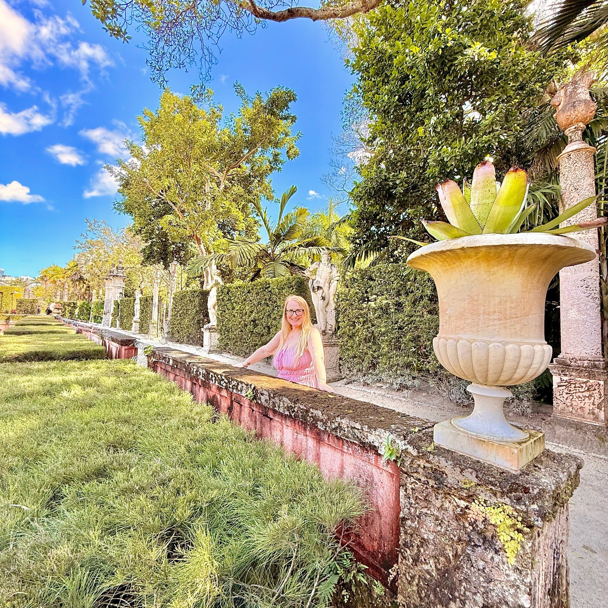 A blonde woman in a pink dress is smiling into the camera. She has her arms on a stone ledge and there is a lush, green garden surrounding her.
