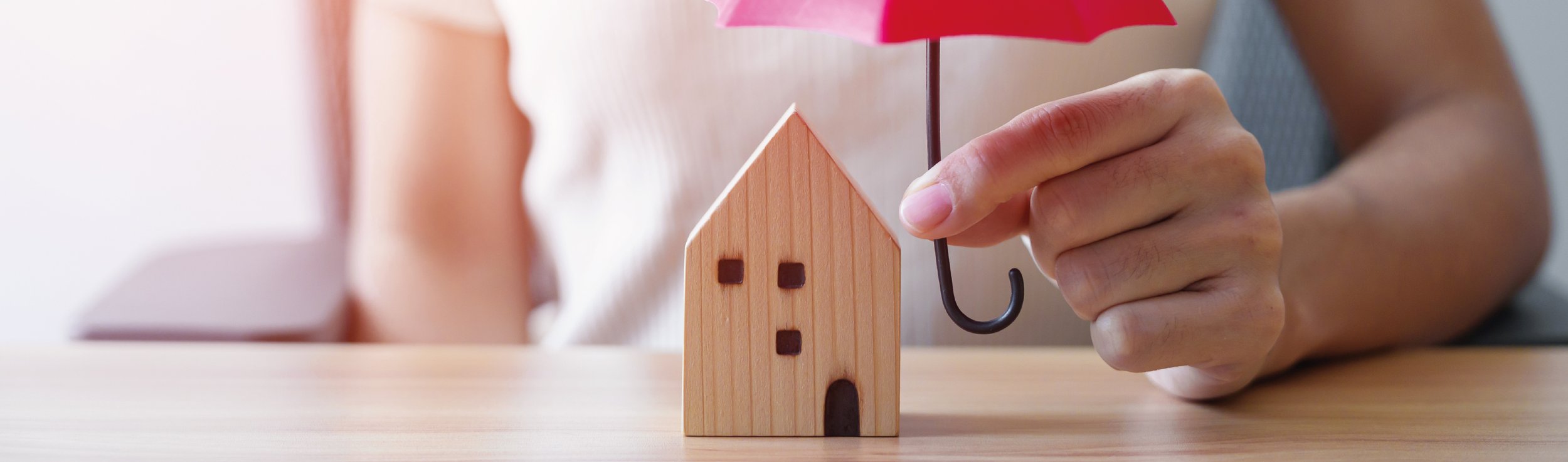 Mão segurando um guarda-chuva rosa acima de uma miniatura de casa de madeira