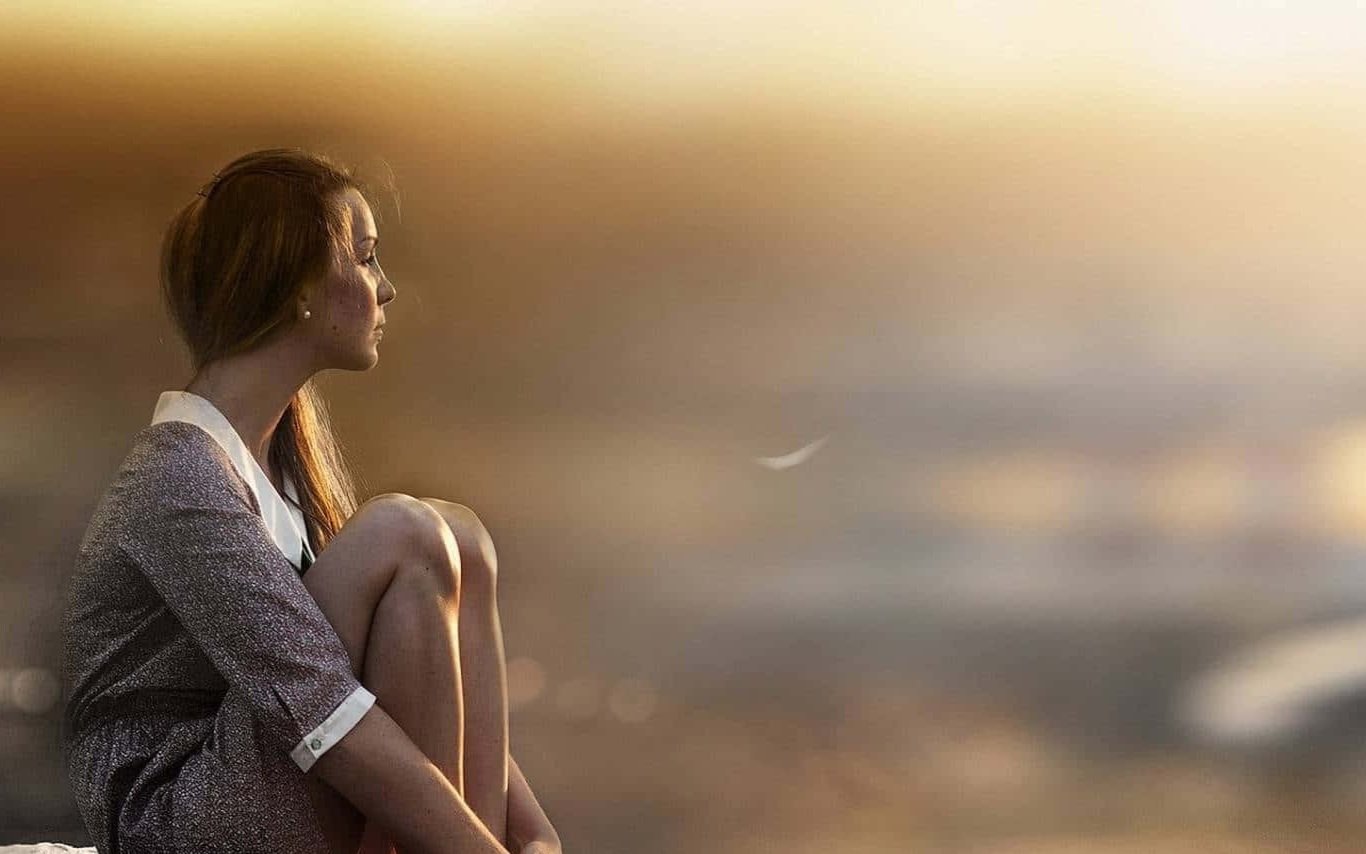 Young woman sitting and looking at sunset, with knees up, in a thoughtful pose.