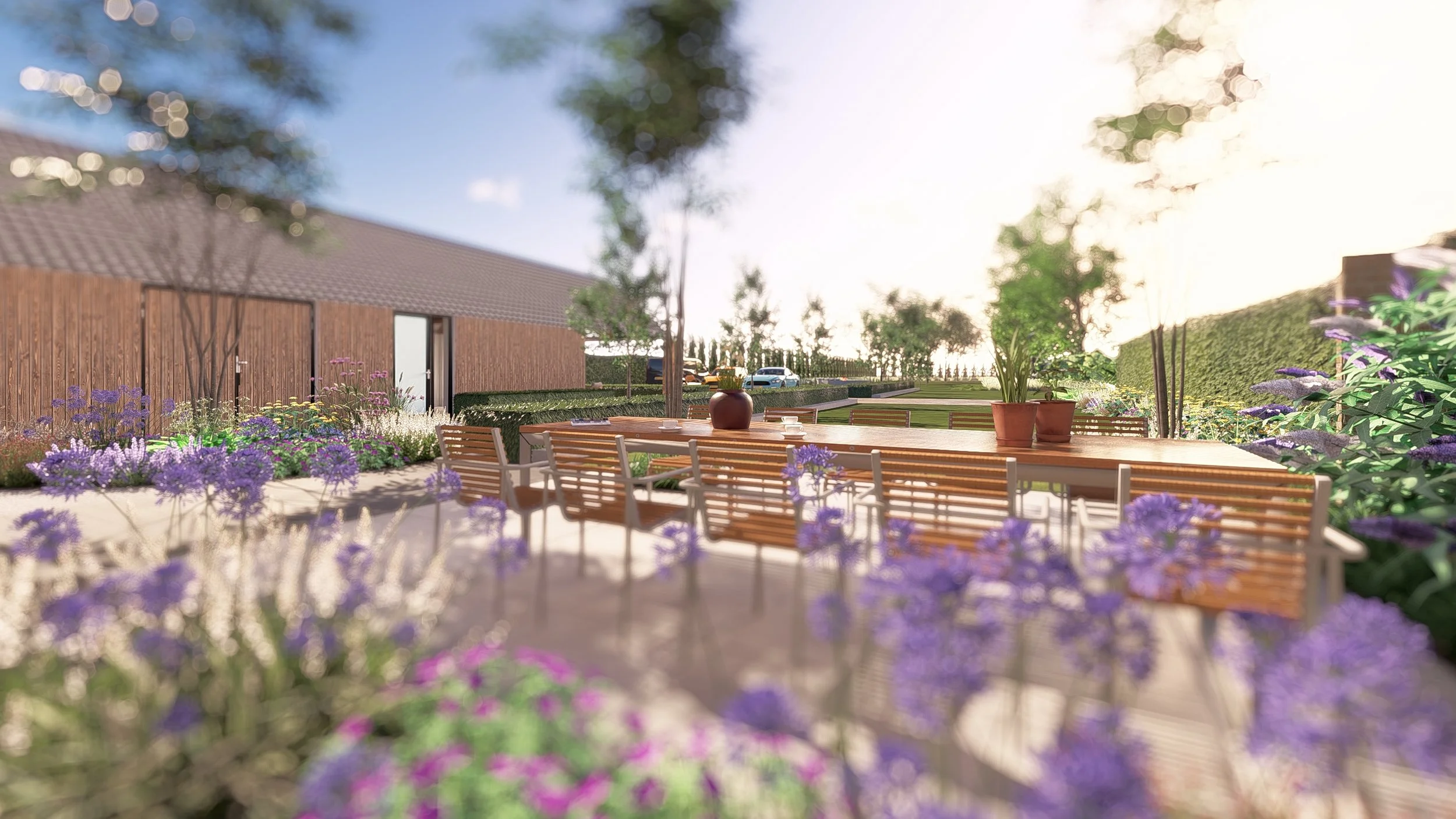 Een tuin met een houten tafel en banken, omringd door bloemen en planten, onder een boom, met een open lucht en een woning ernaast.