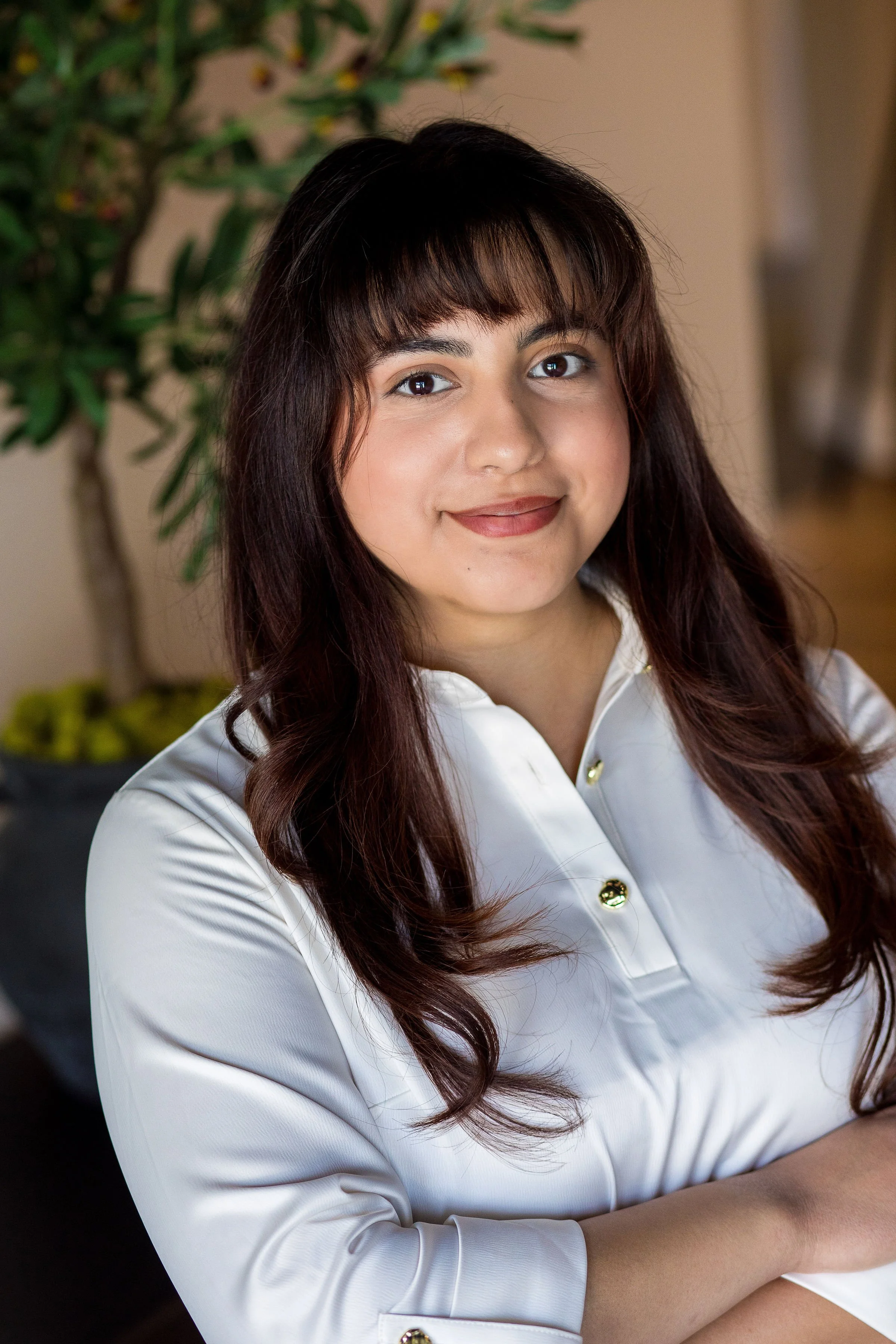 A young woman with long, dark brown hair and bangs smiling while wearing a white shirt with gold buttons, standing in front of a blurred indoor background with a plant.