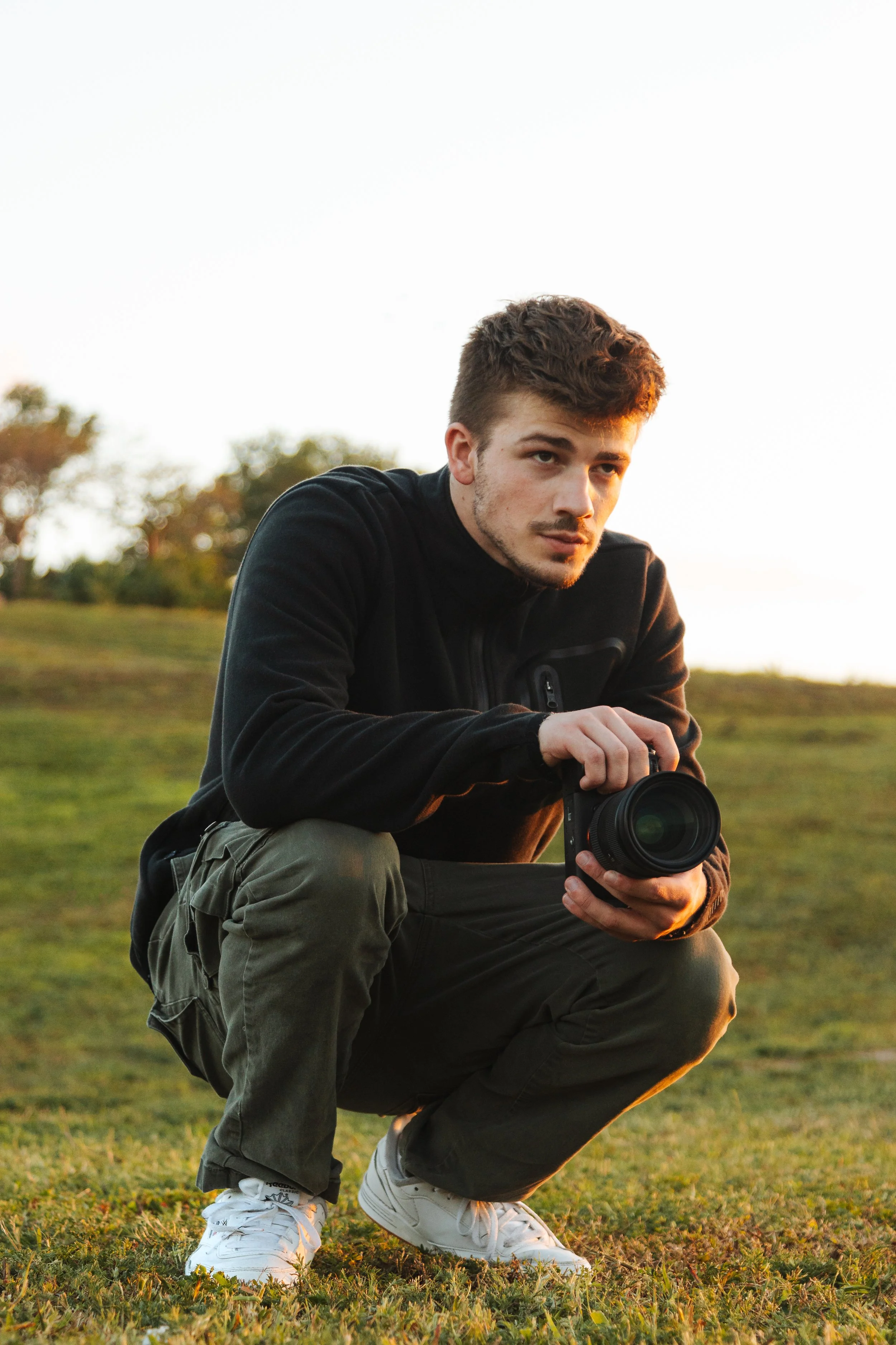 A young man with short brown hair, crouching outdoors on grass, holding a camera, during sunset.