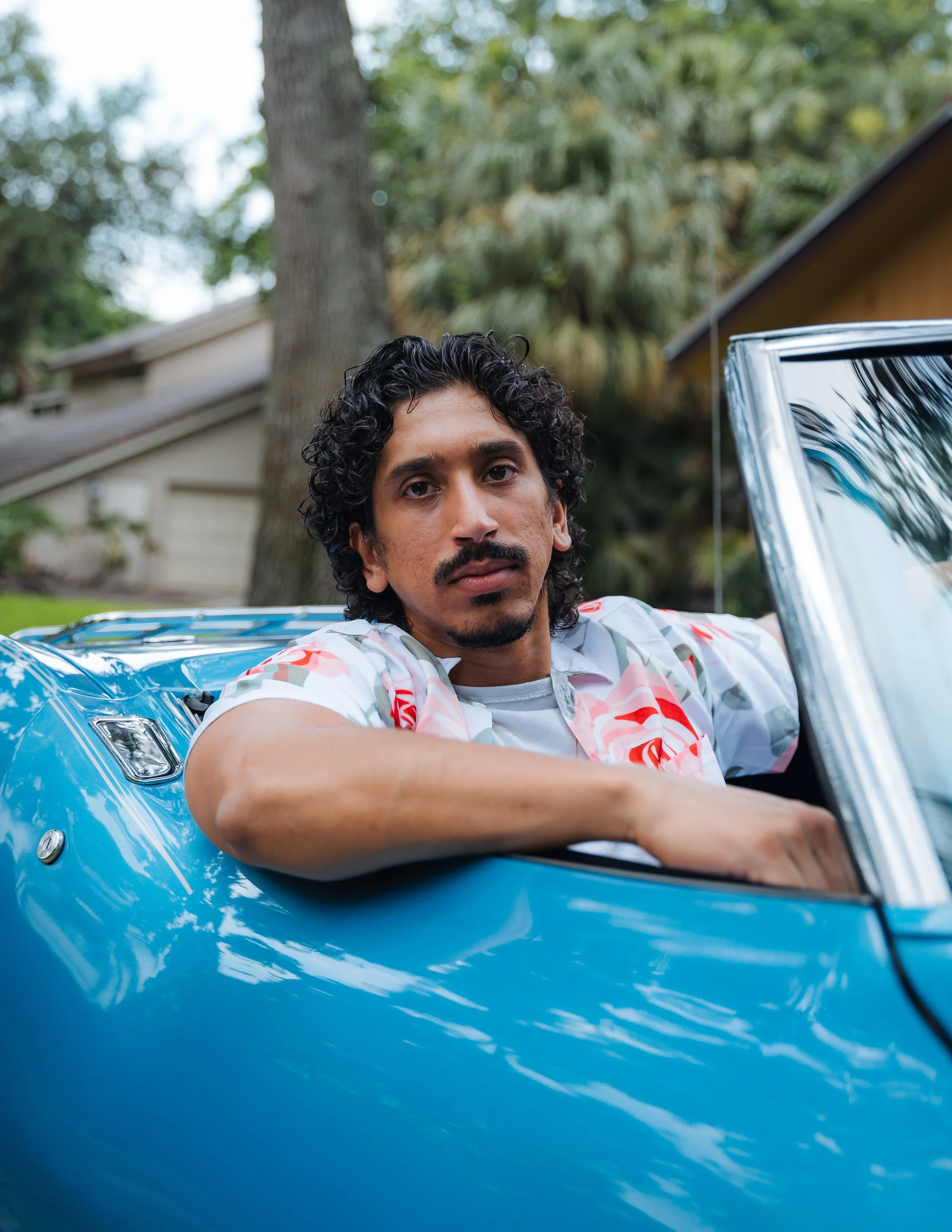 A man with curly black hair, a mustache, and a goatee sitting in a blue convertible car with the door open, looking at the camera. There are trees and a house in the background.