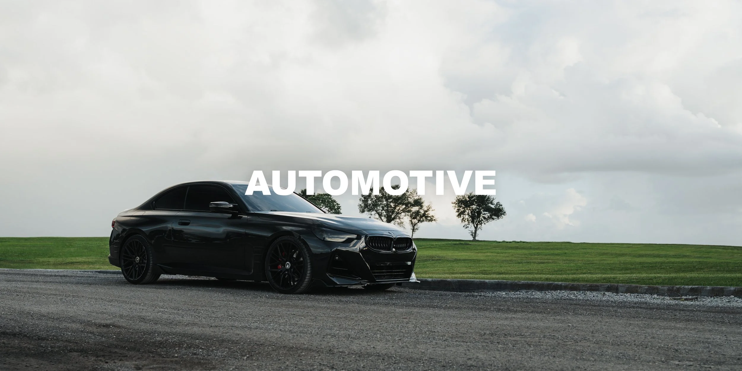 Black BMW parked on a road with a green field and trees in the background under a cloudy sky