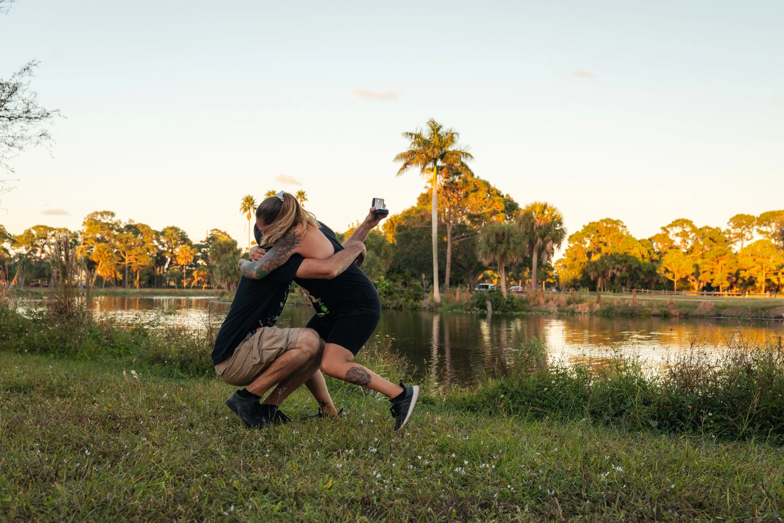A couple hugging beside a lake during sunset, with palm trees and lush greenery in the background.