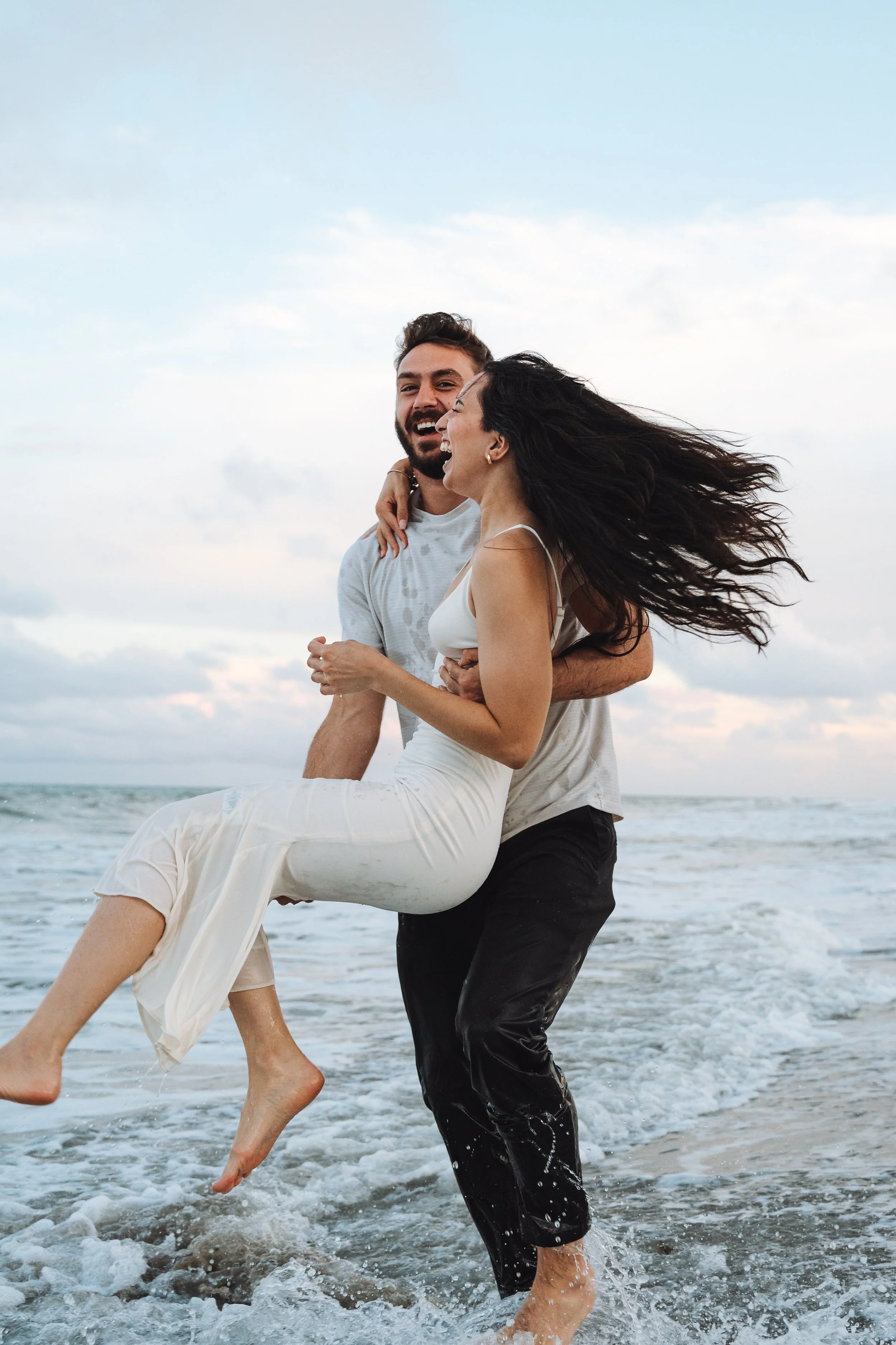 A couple enjoying a playful moment in the ocean, with the man lifting the woman as they laugh near the shoreline.