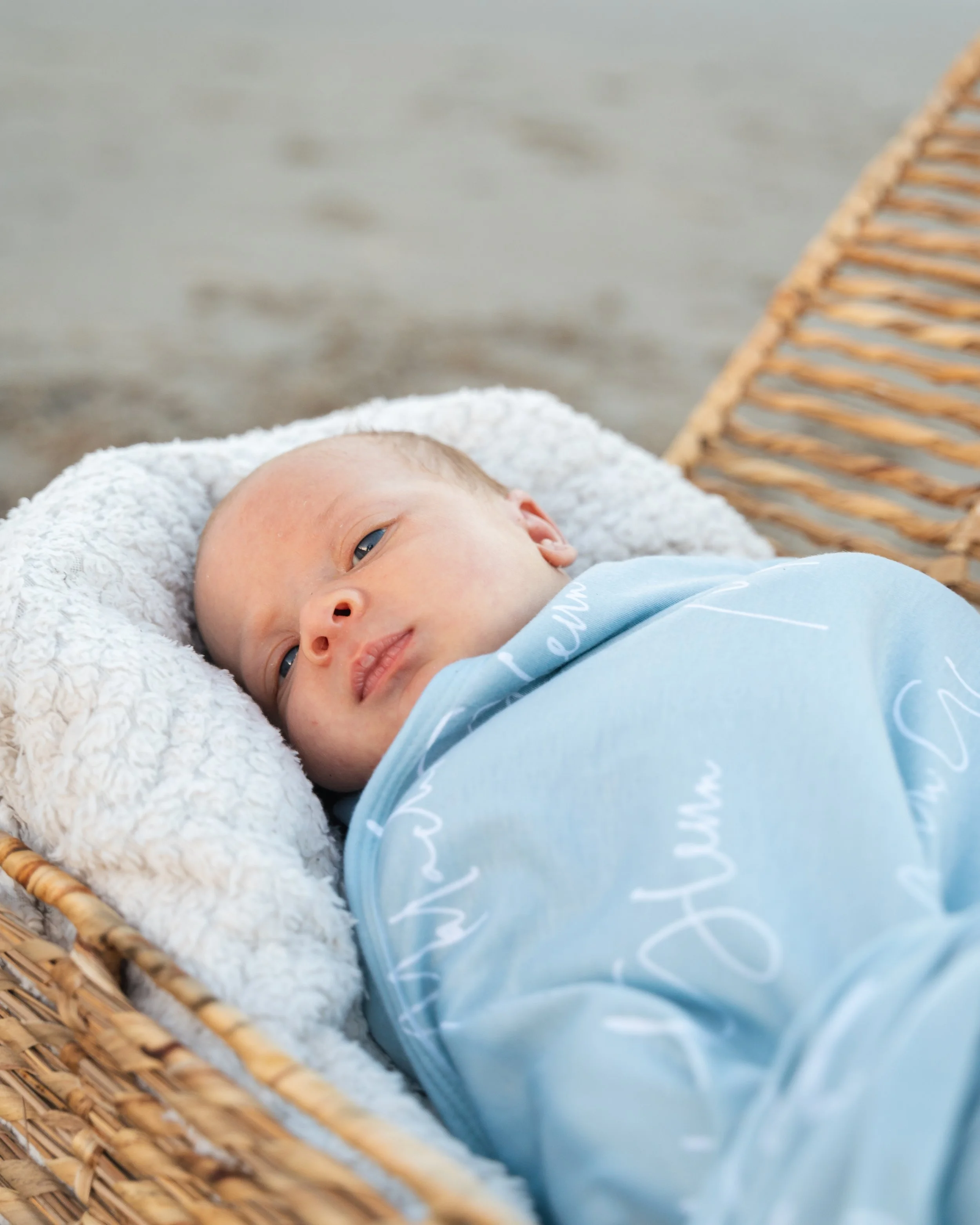 Baby lying in a wicker basket on a soft blanket, wearing a blue outfit, with a blurred beach background.