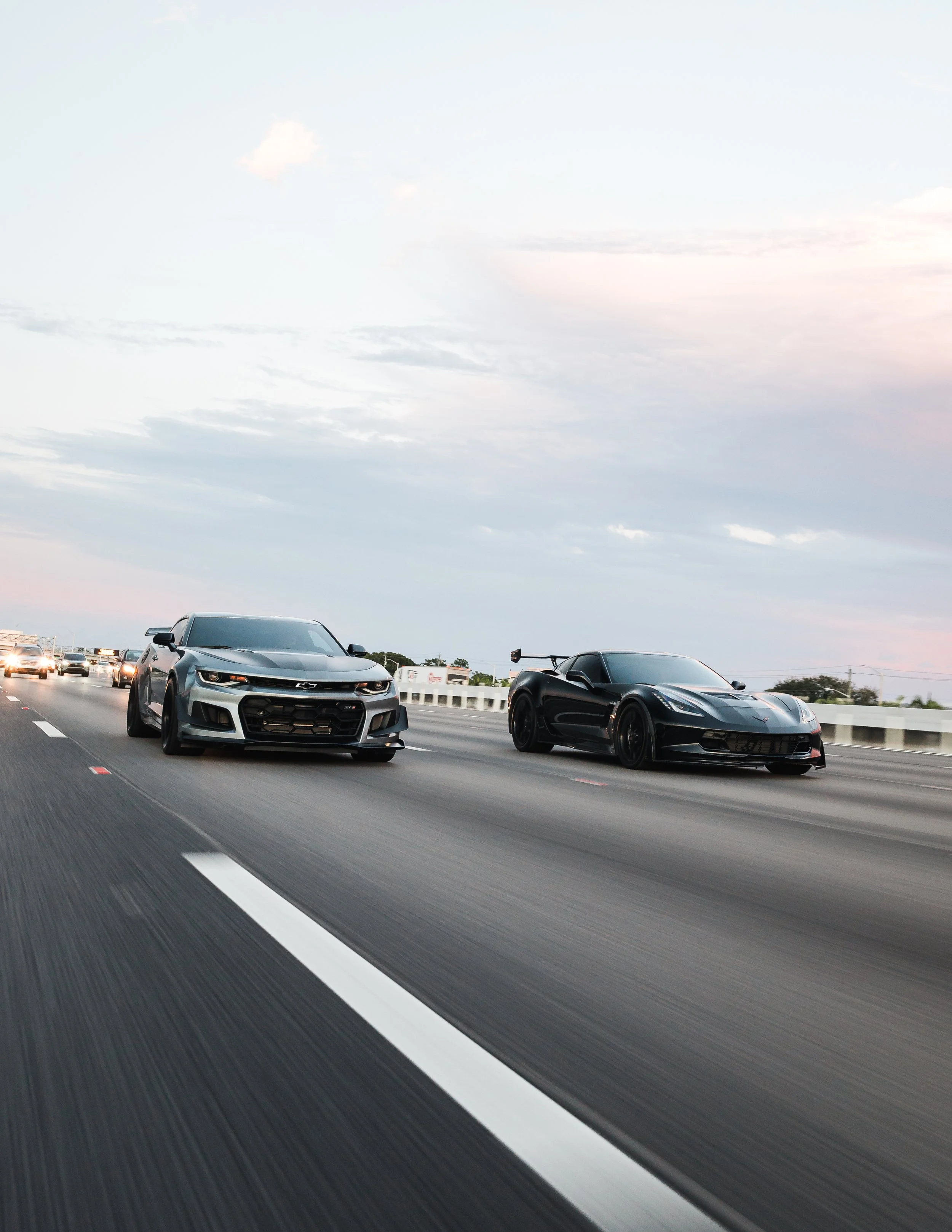 Two sports cars racing on a highway during dusk, one black and one silver, with traffic in the background.