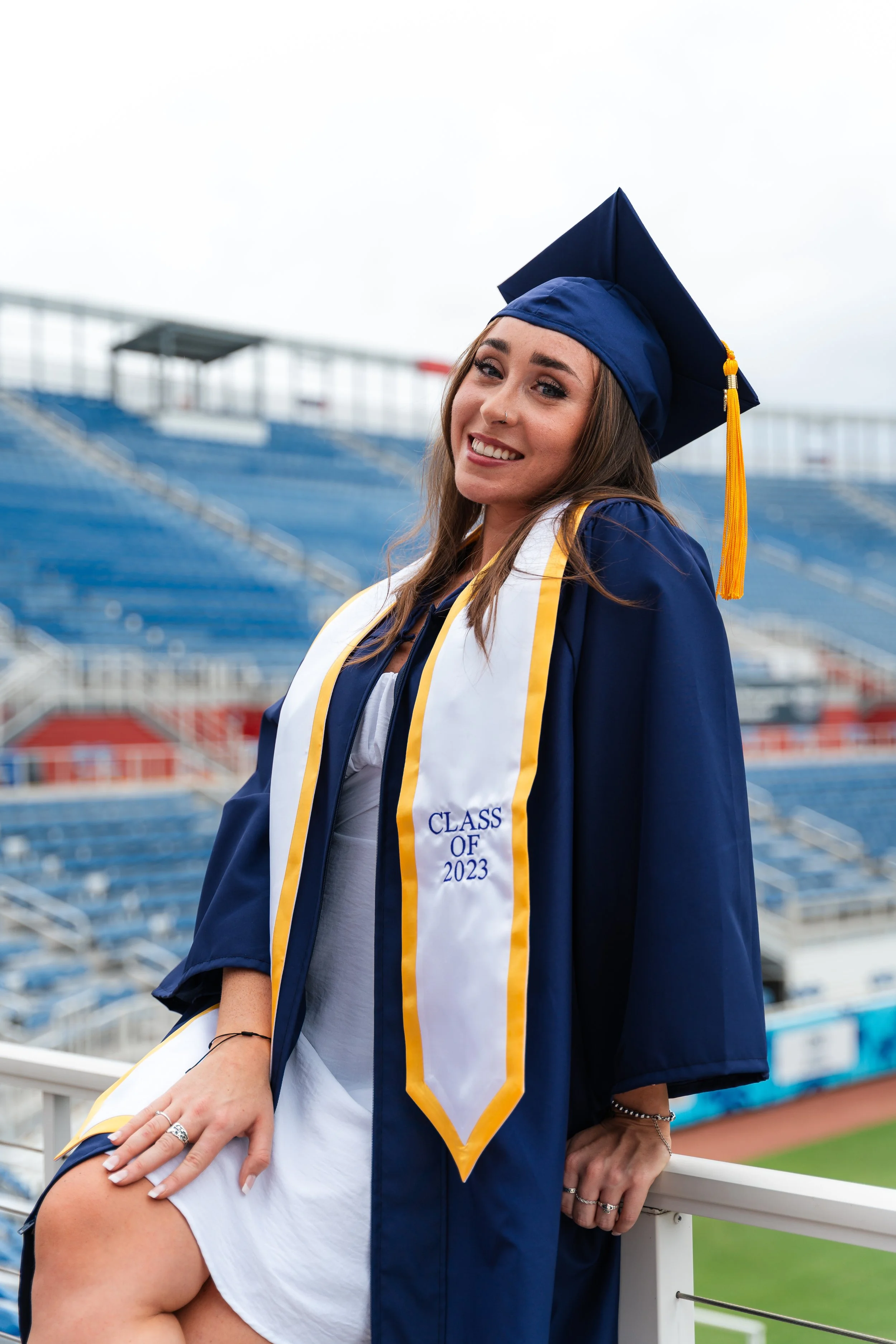 A young woman in a blue graduation cap and gown posing at a stadium, wearing a white sash with yellow trim that says 'Class of 2023'.