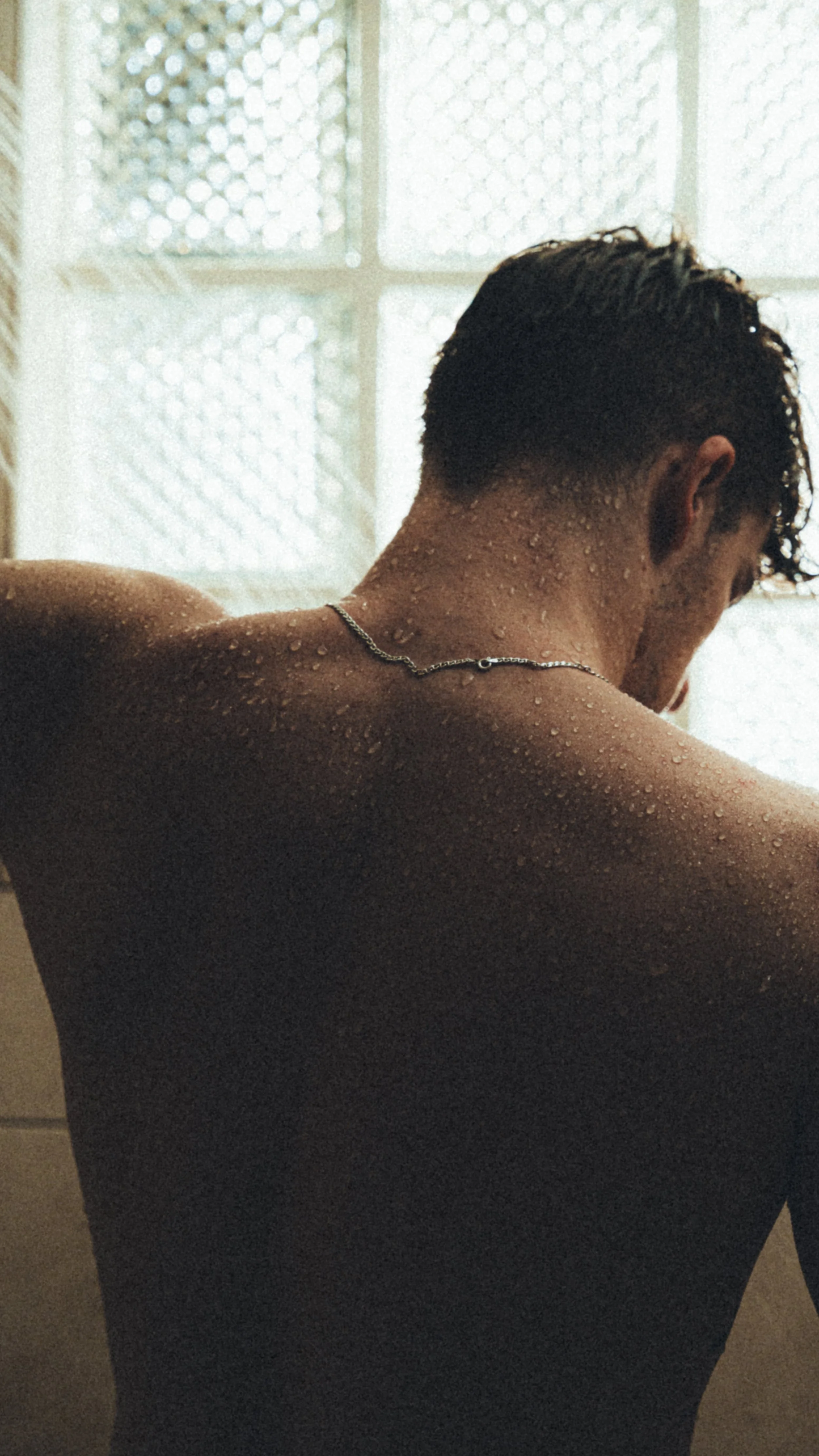 A man with wet skin and hair, seen from behind, sitting in front of a frosted window with patterned glass, wearing a chain necklace.