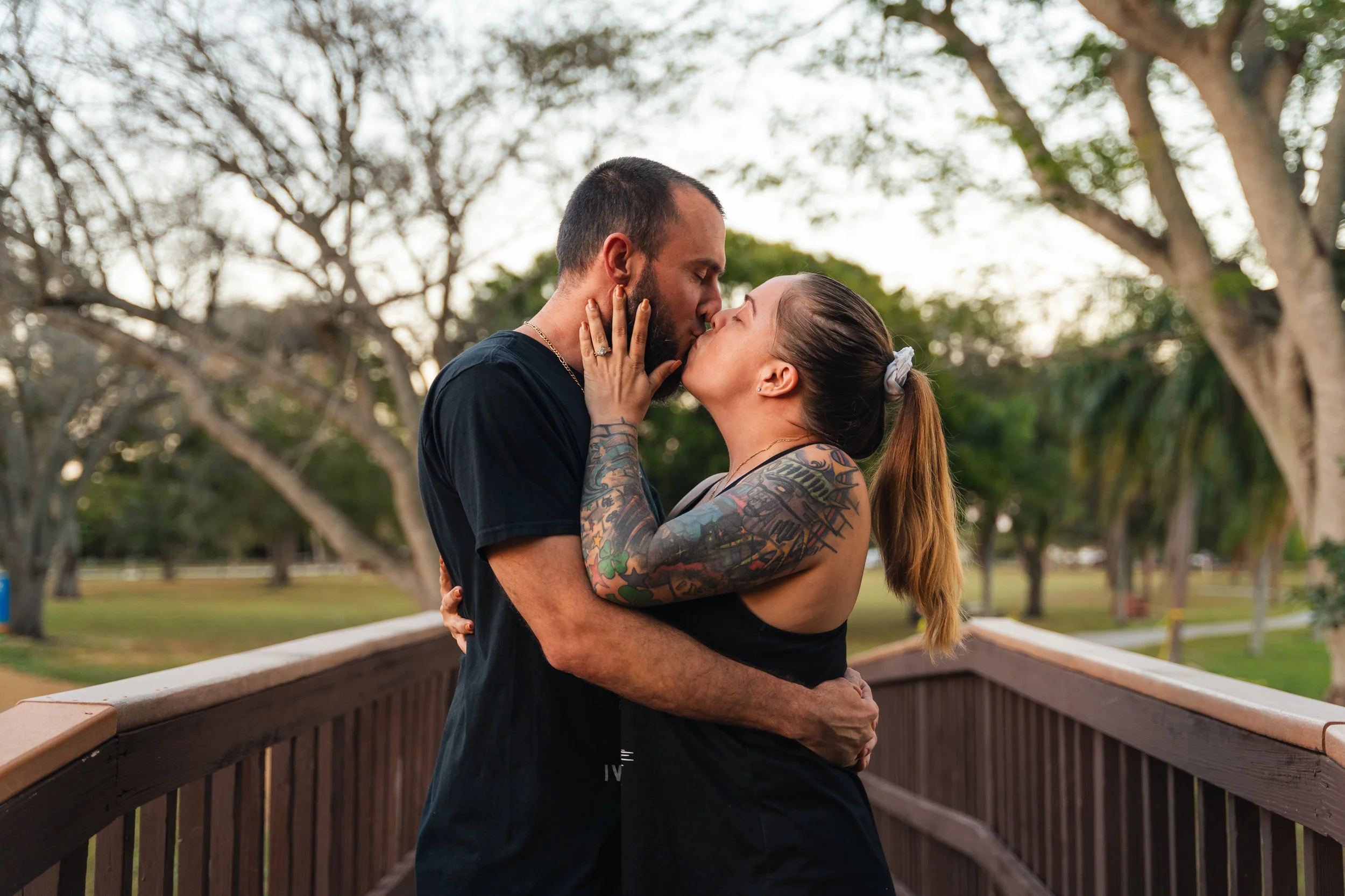 A couple sharing a kiss outdoors on a wooden bridge, with trees and a park in the background.