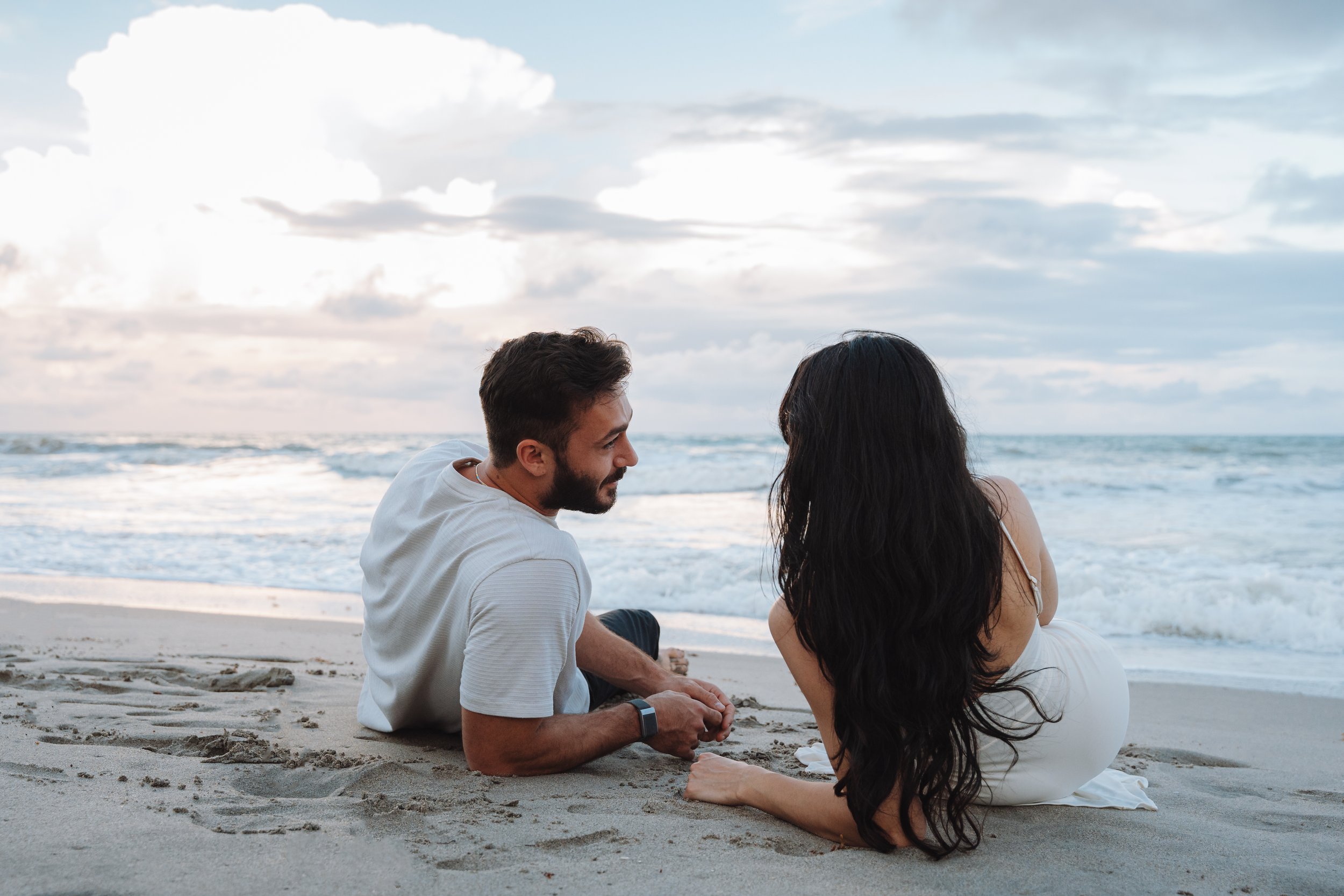 A man and woman sitting on the beach, facing each other, with ocean waves and a cloudy sky in the background.