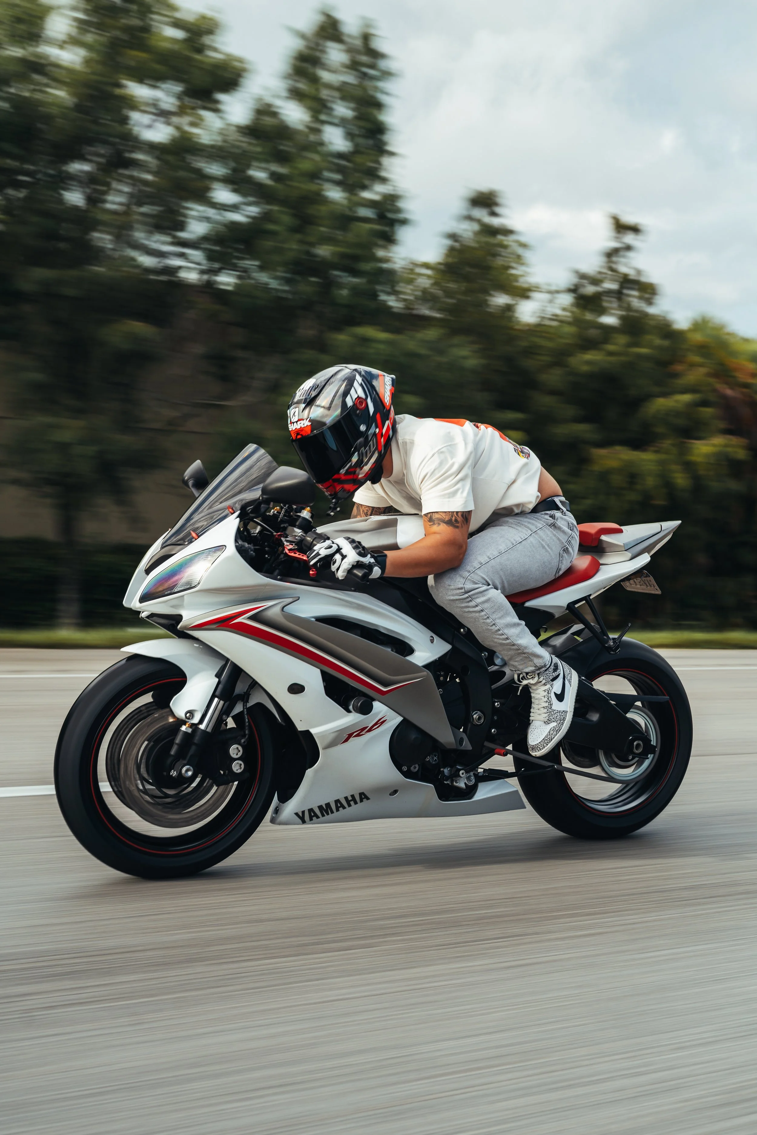 A person riding a Yamaha sport motorcycle at high speed on a road, wearing a helmet, white t-shirt, gray pants, and sneakers, with motion blur in the background.