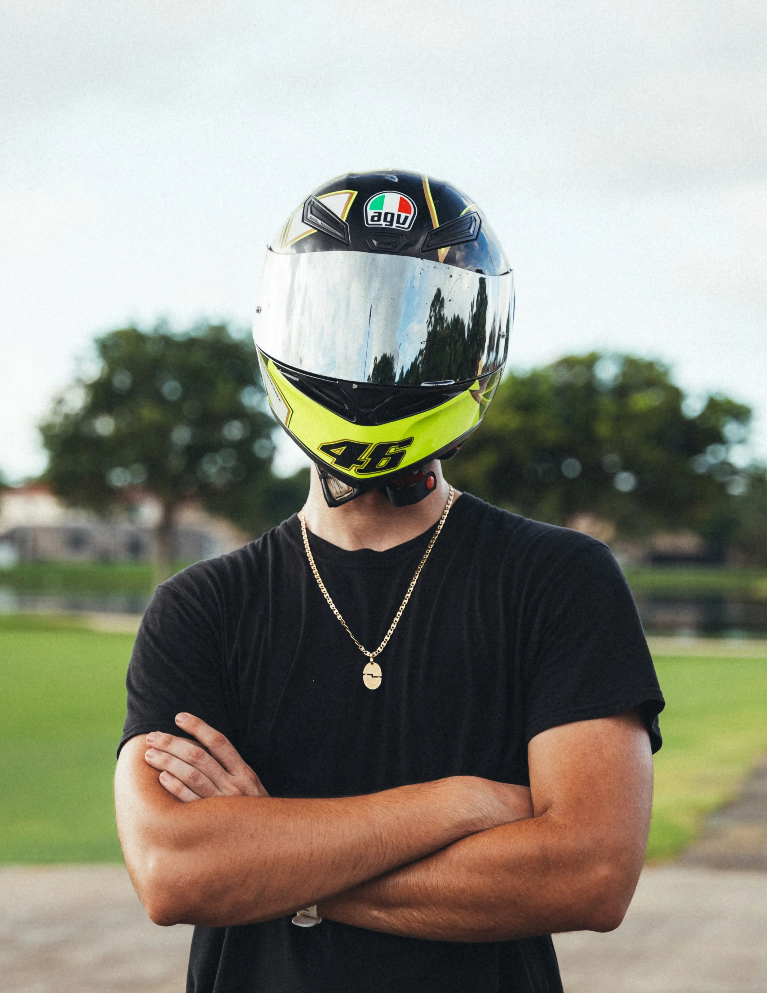 Person standing outdoors with arms crossed, wearing a black t-shirt, a gold chain necklace, and a full-face motorcycle helmet with a reflective visor, black and yellow accents
