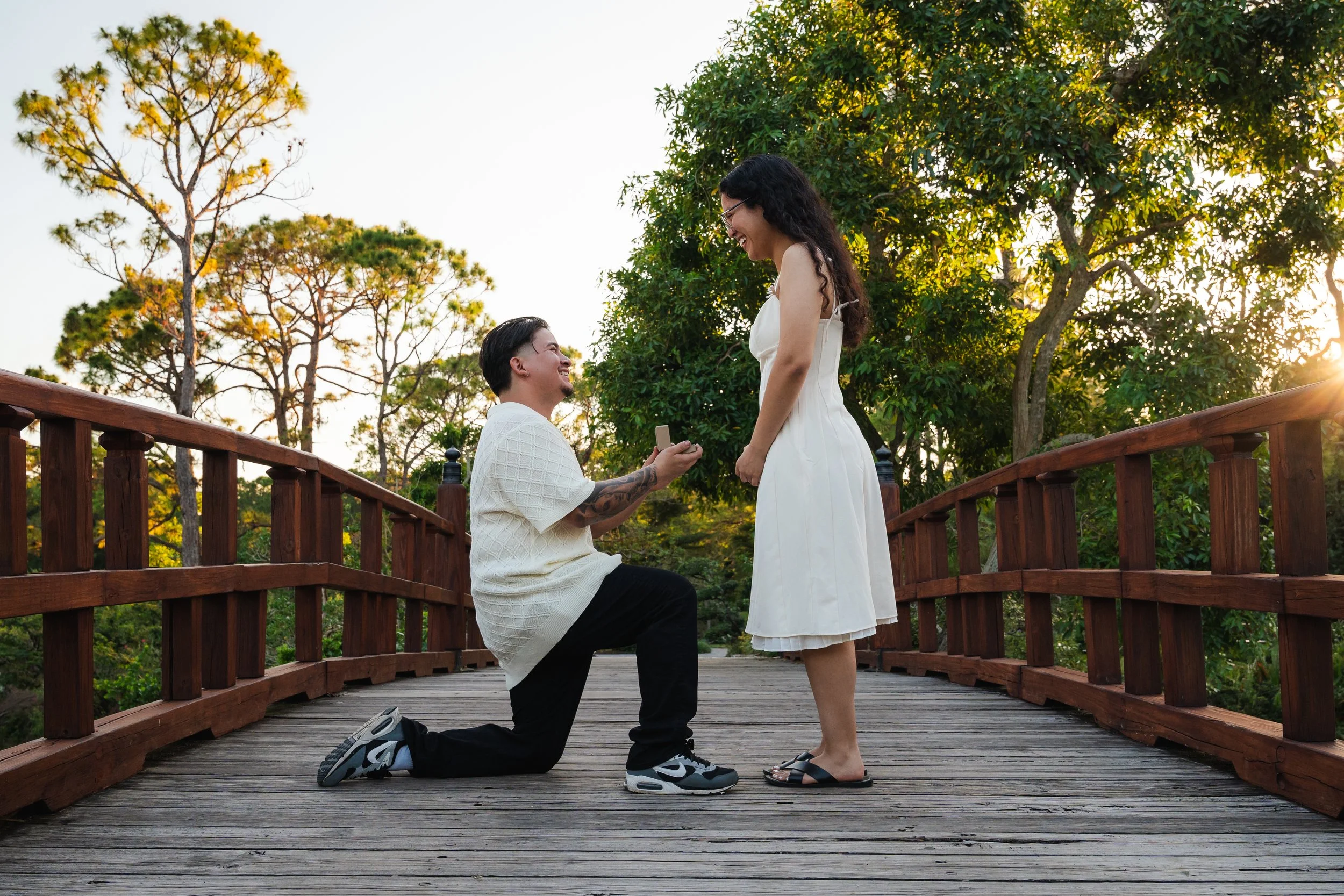 A man proposing marriage to a woman on a wooden bridge in a park during sunset, with trees in the background.