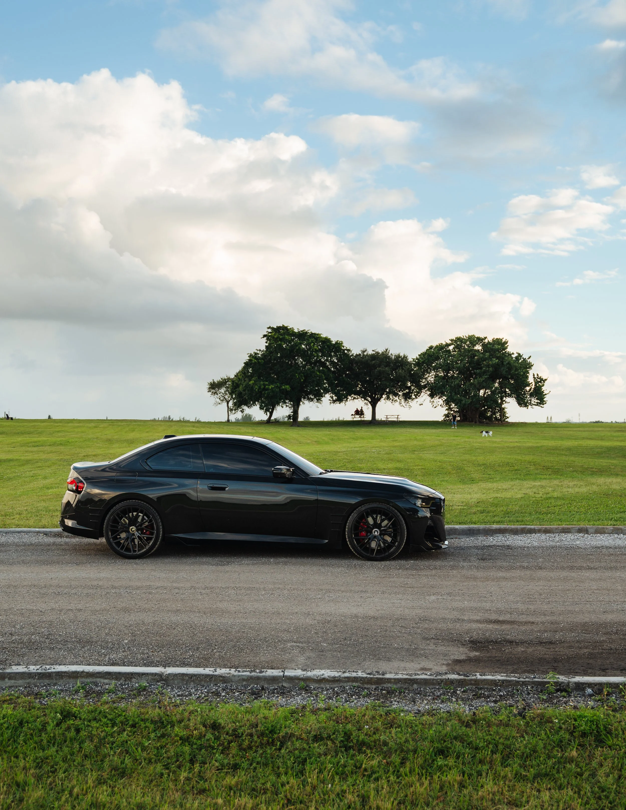A black sports car parked on a gravel road, with a grassy field, trees, and a cloudy sky in the background.