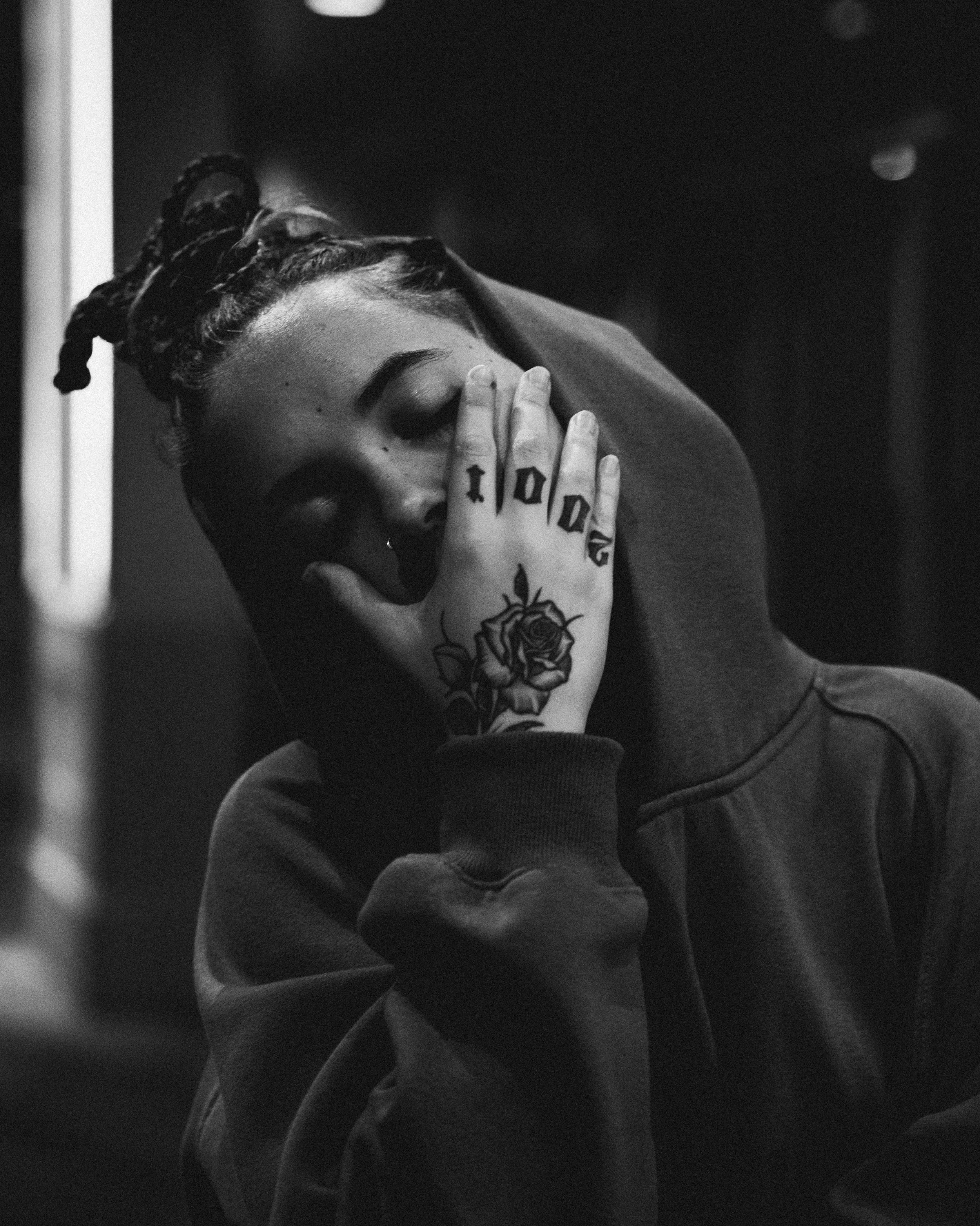 Black and white photo of a person with dreadlocks covering part of their face, resting their head on their hand with tattoos on fingers and a rose on the hand, wearing a hoodie.