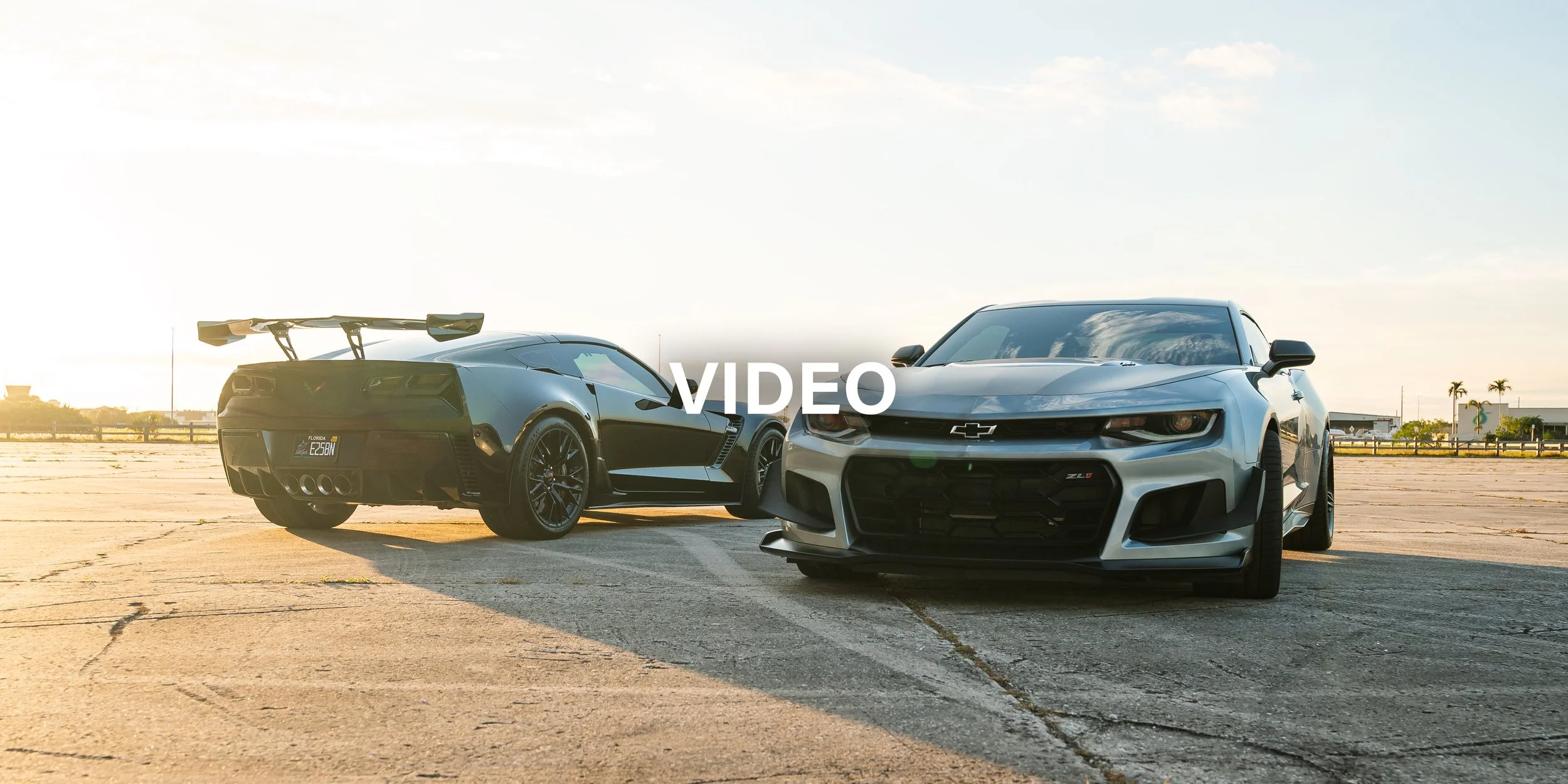 Two Chevrolet sports cars, one black with a large rear spoiler and one gray, are parked on an empty concrete lot during sunset