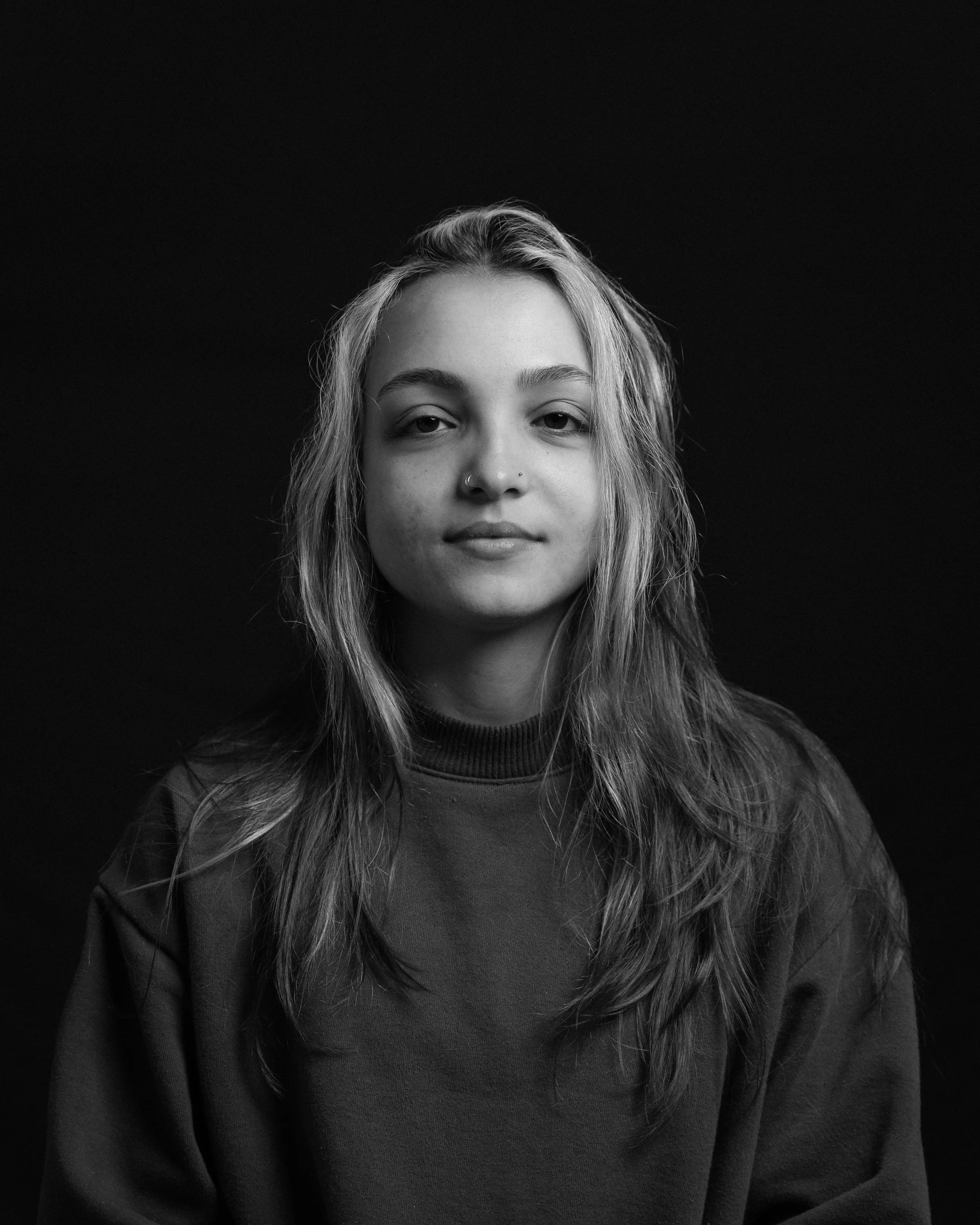Black and white portrait of a young woman with long hair, wearing a dark sweatshirt, looking directly at the camera against a dark background.