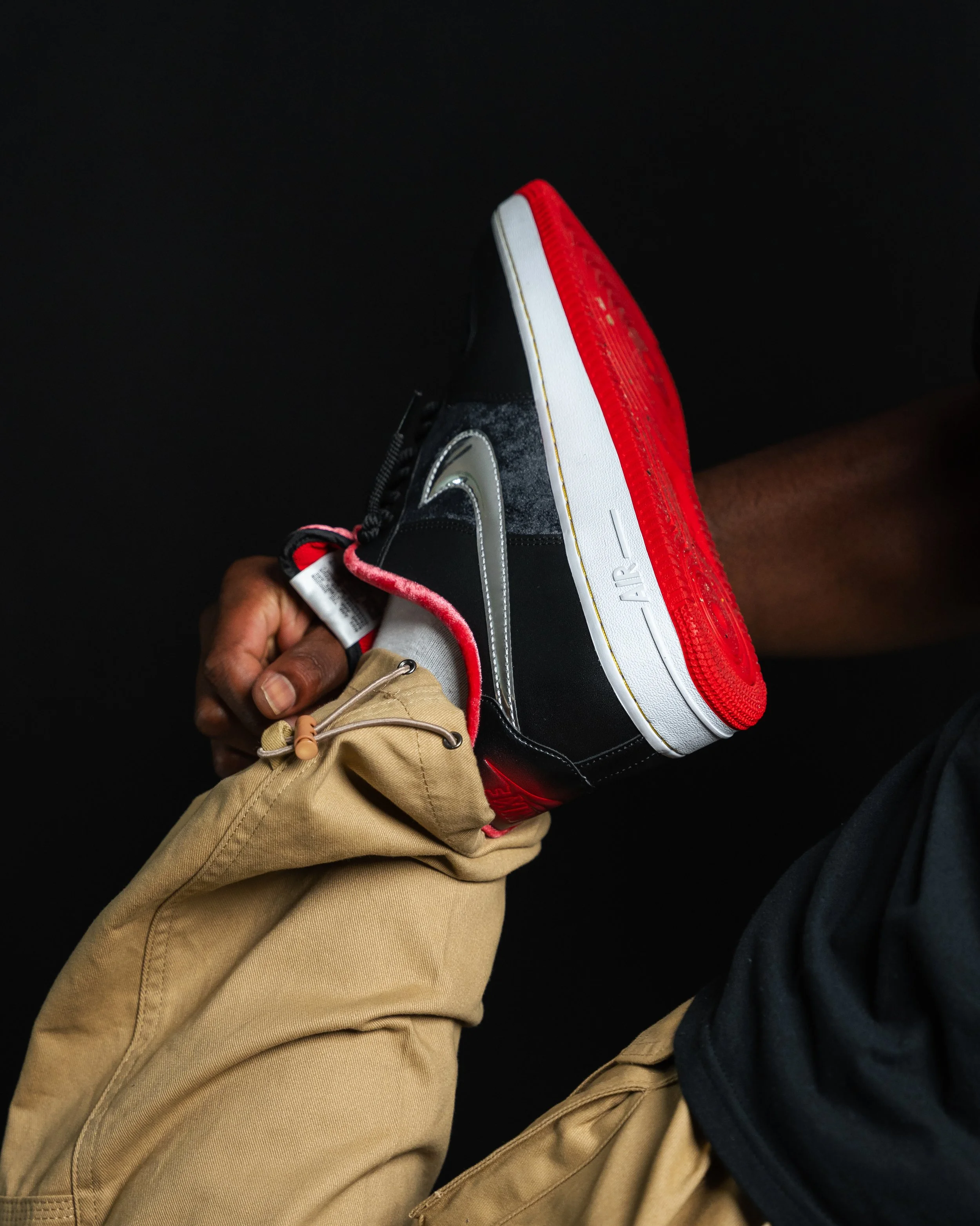 A person wearing tan pants and a black shirt holding a black, white, and red Nike Air Jordan sneaker against a black background.