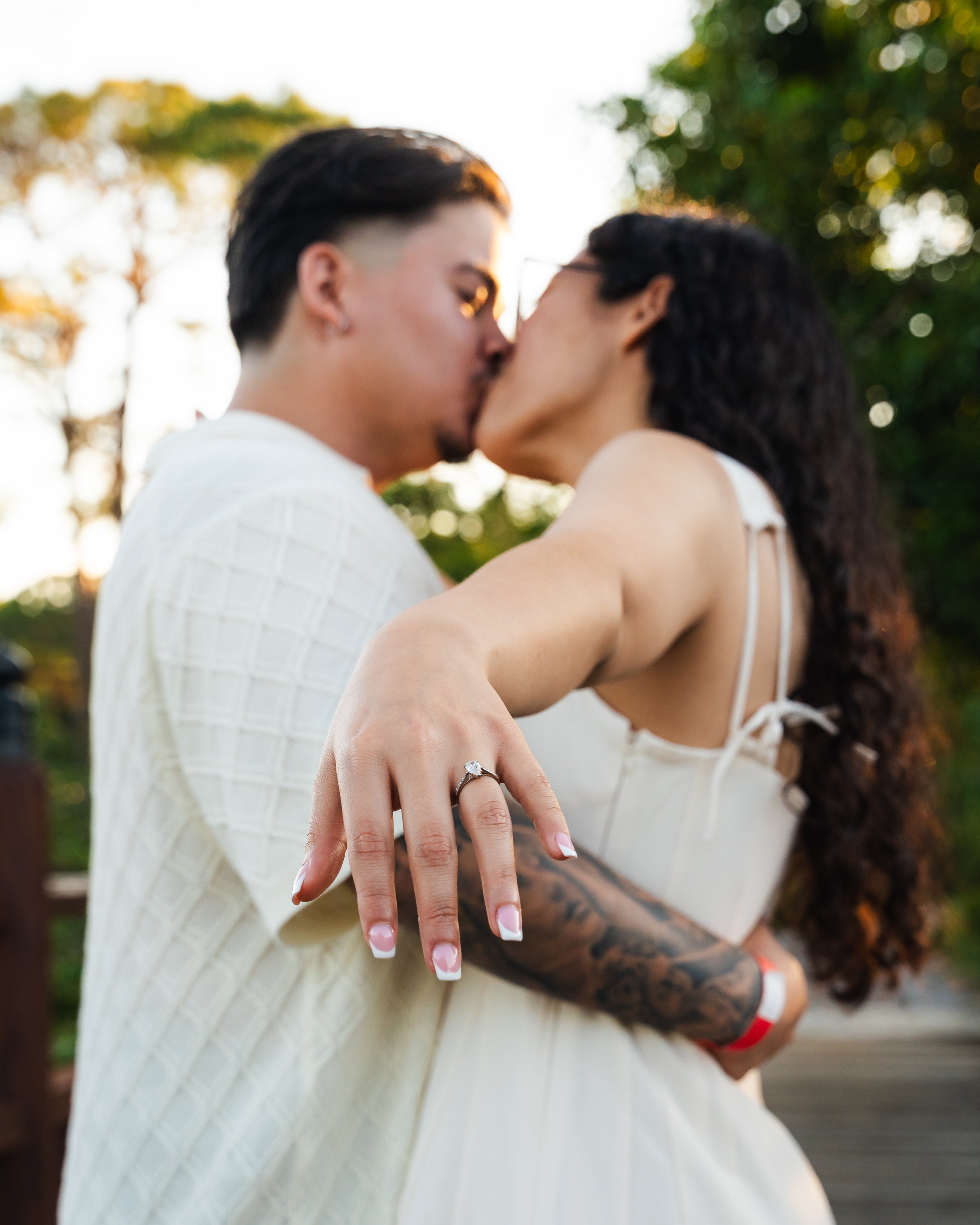 A couple sharing a kiss outdoors at sunset, with the woman showing off her engagement ring on her left hand.