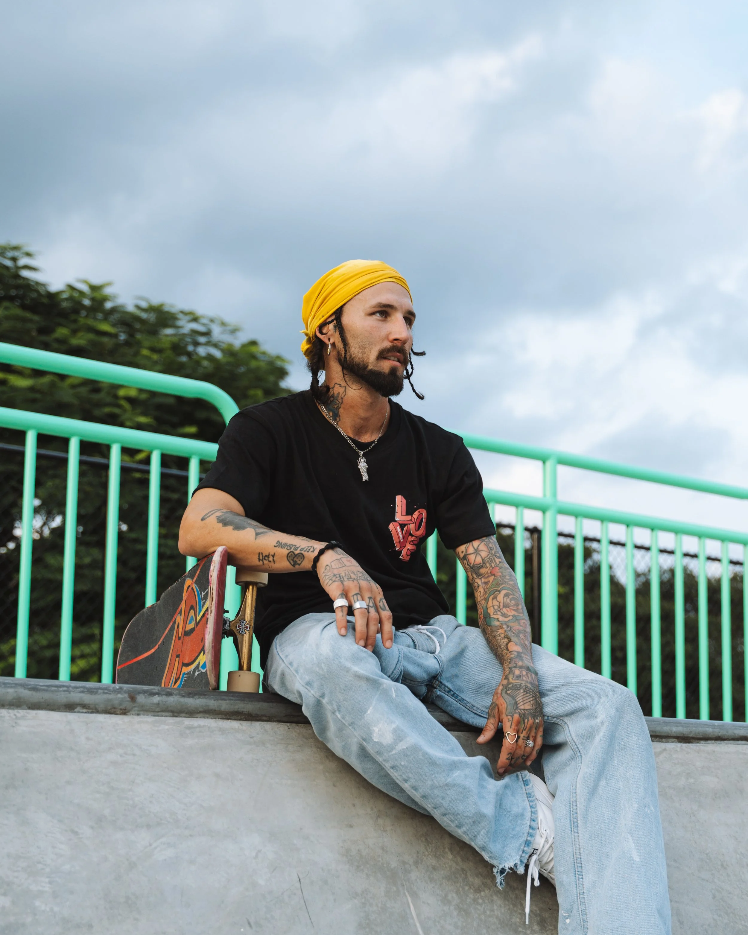 A young man with tattoos, a yellow headscarf, black t-shirt, and ripped jeans sitting on a skate ramp with a skateboard next to him, under a cloudy sky.