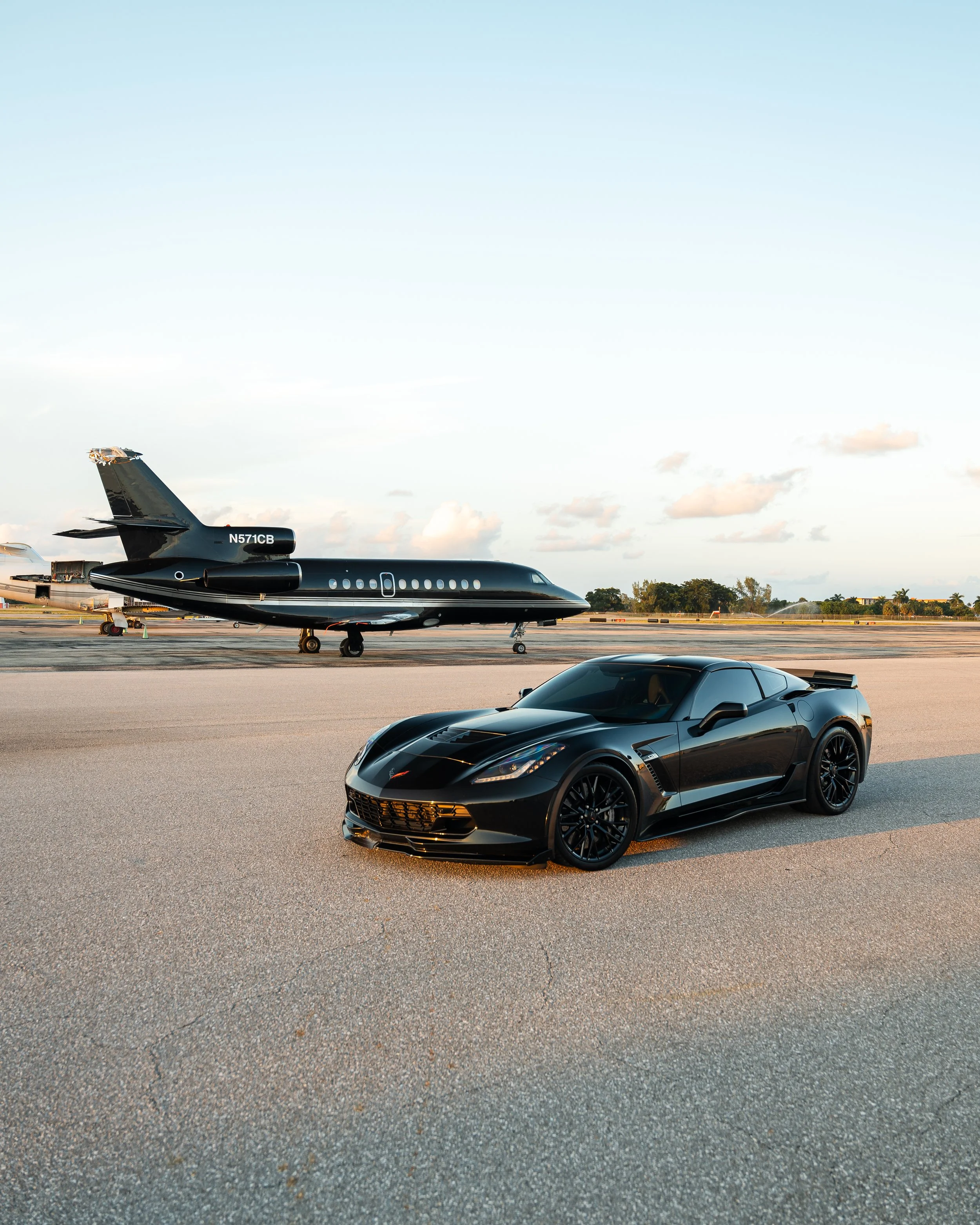 Black sports car parked on tarmac with a private jet in the background at an airport during sunset.