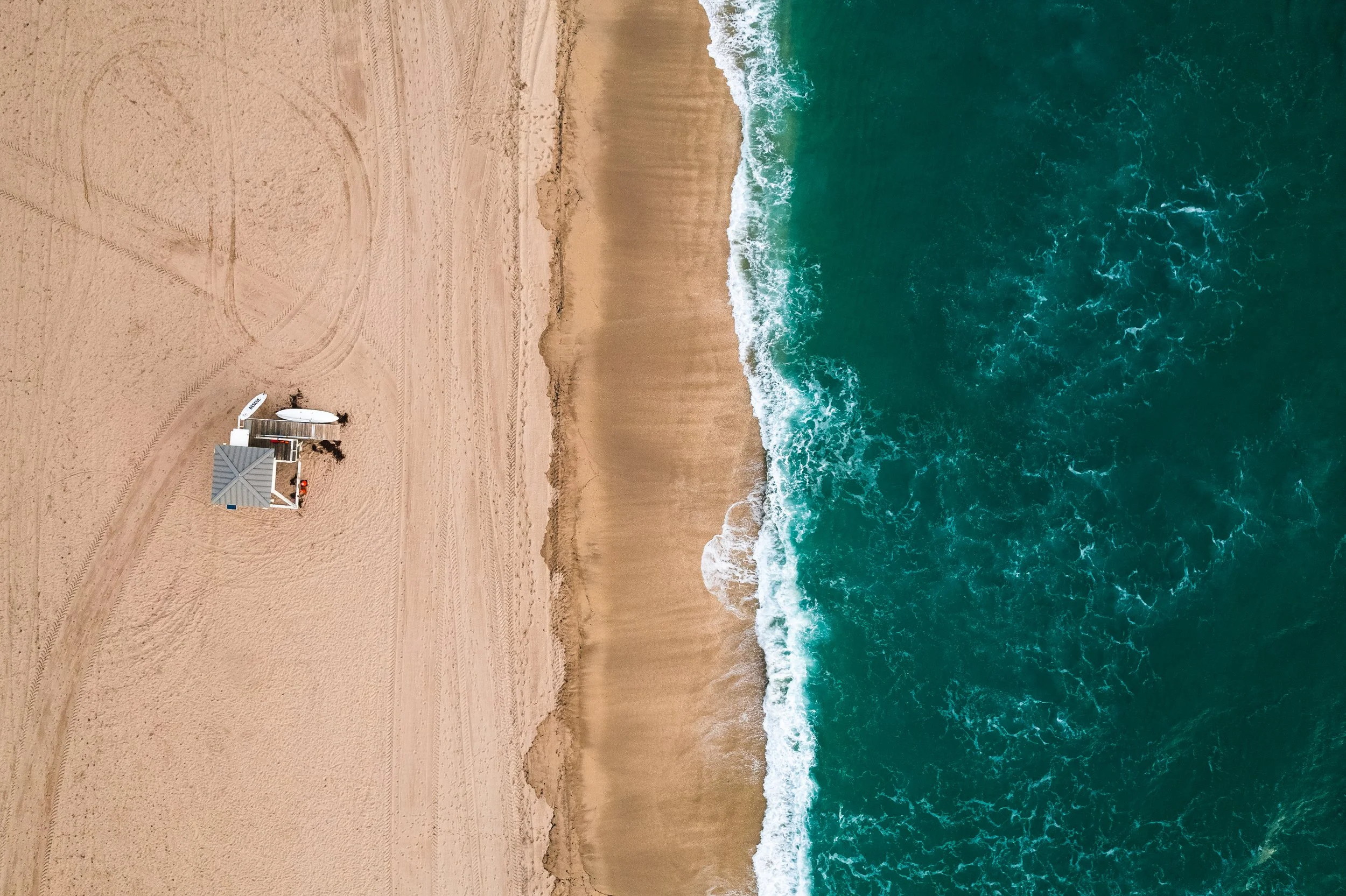 Aerial view of a sandy beach with a surfboard and a small shaded structure near the shoreline, ocean waves crashing onto the beach.
