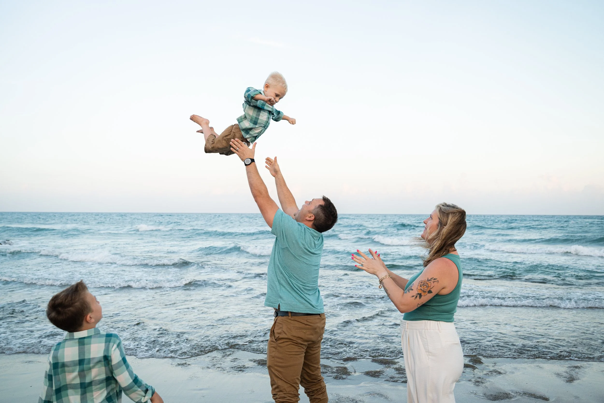 Family of four at the beach, with a man lifting a young boy into the air, the boy smiling, woman clapping, and an older boy watching on, ocean in the background.