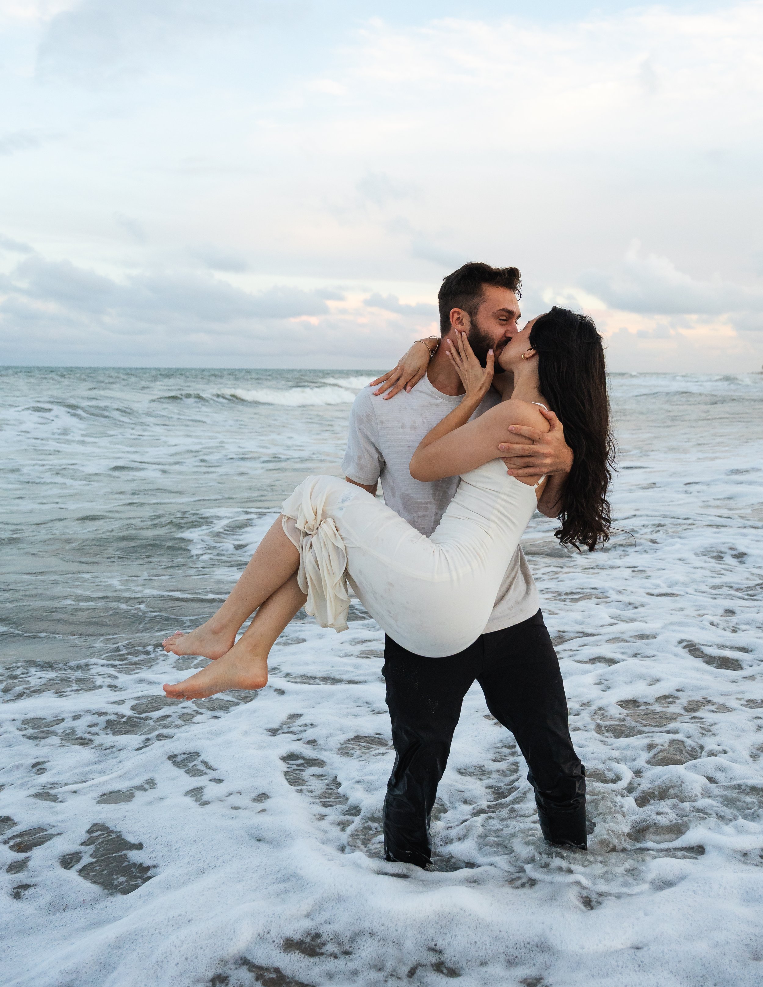 A couple sharing a kiss in the ocean, with the man holding the woman in his arms as waves wash around their feet.