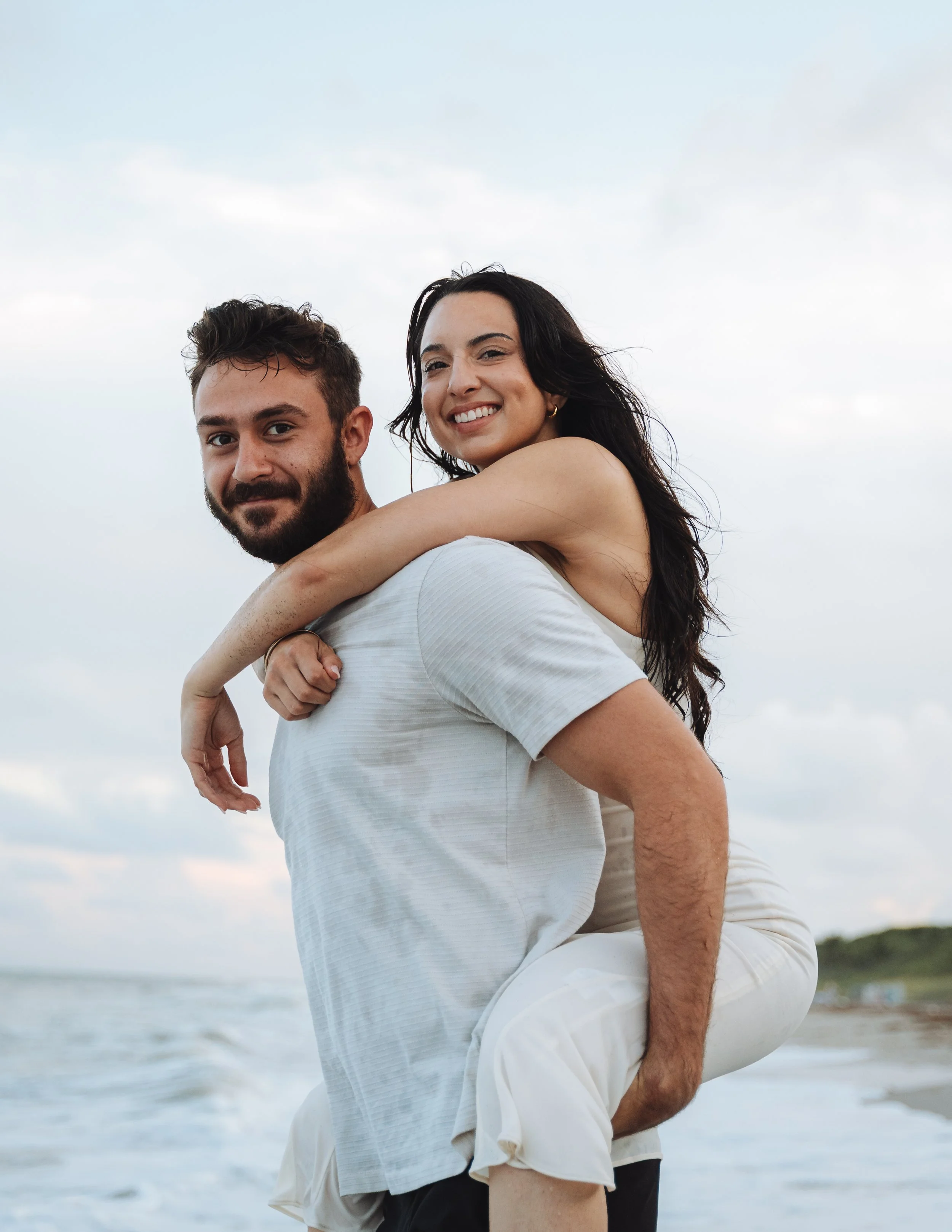 A smiling woman is riding on a man's back along the beach, with the ocean and sky in the background.