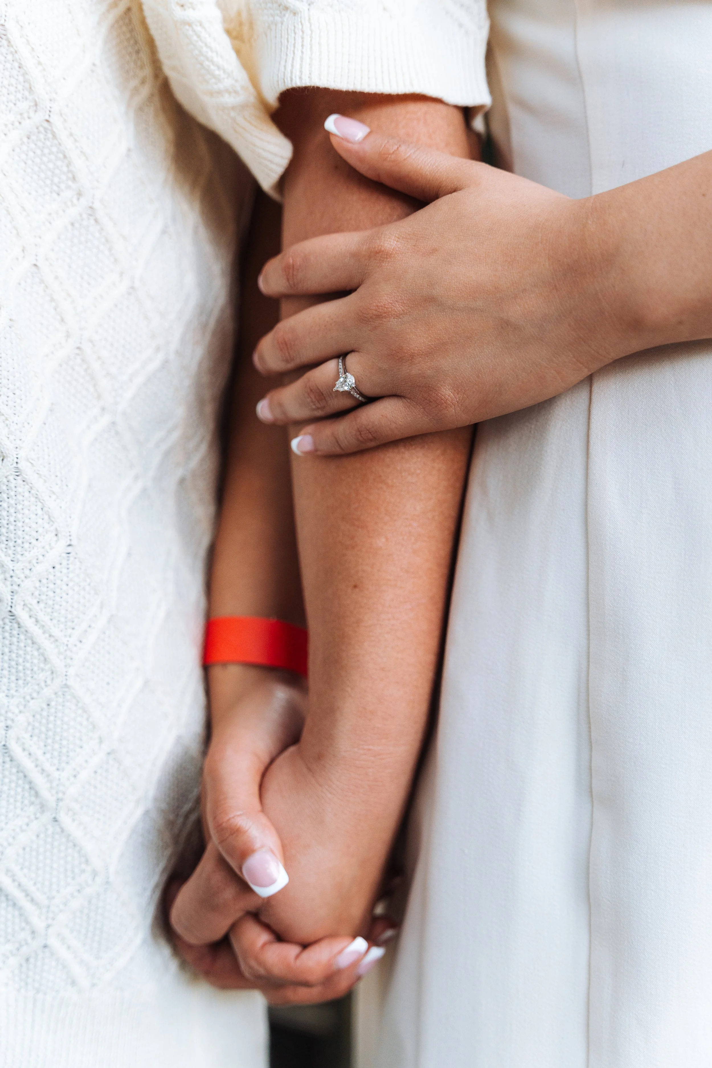 Close-up of two people holding hands; one is wearing a diamond engagement ring, and the other has a red wristband.
