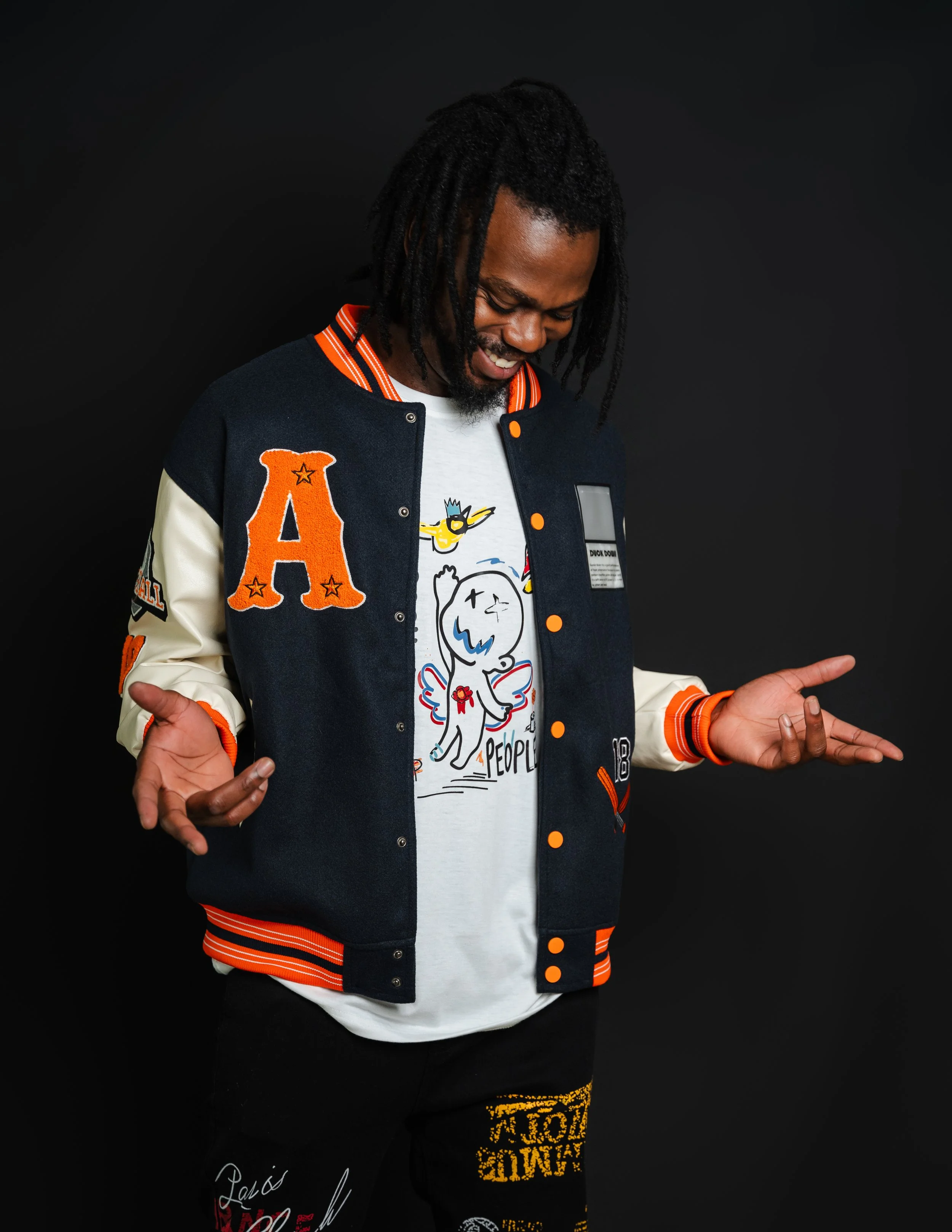 A man with dreadlocks smiling and looking down, wearing a navy and cream letterman jacket with orange accents, and a white graphic T-shirt, standing against a black background.