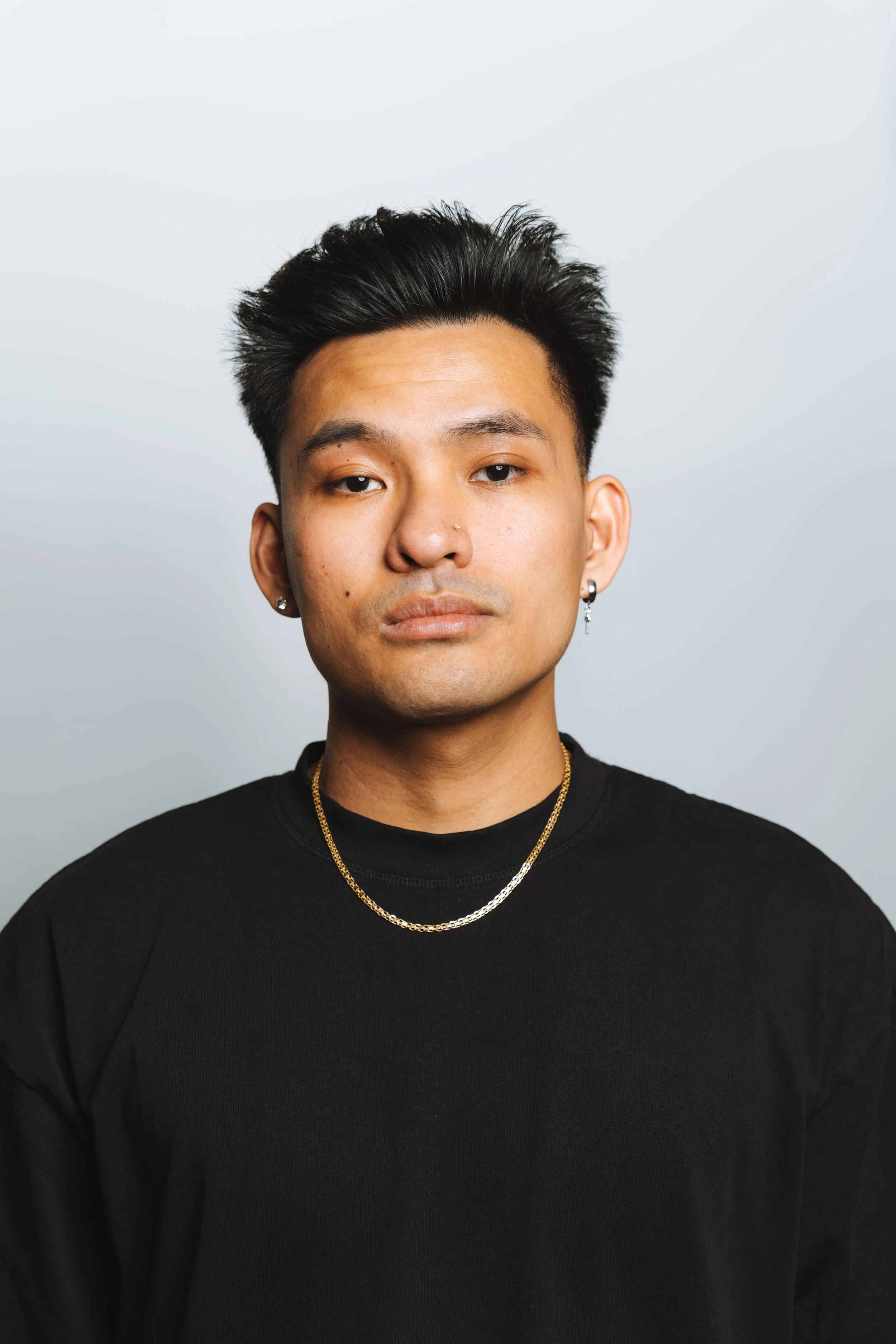 Portrait of a young Asian man with short dark hair, wearing earrings, a nose piercing, and gold necklaces, against a plain light gray background.