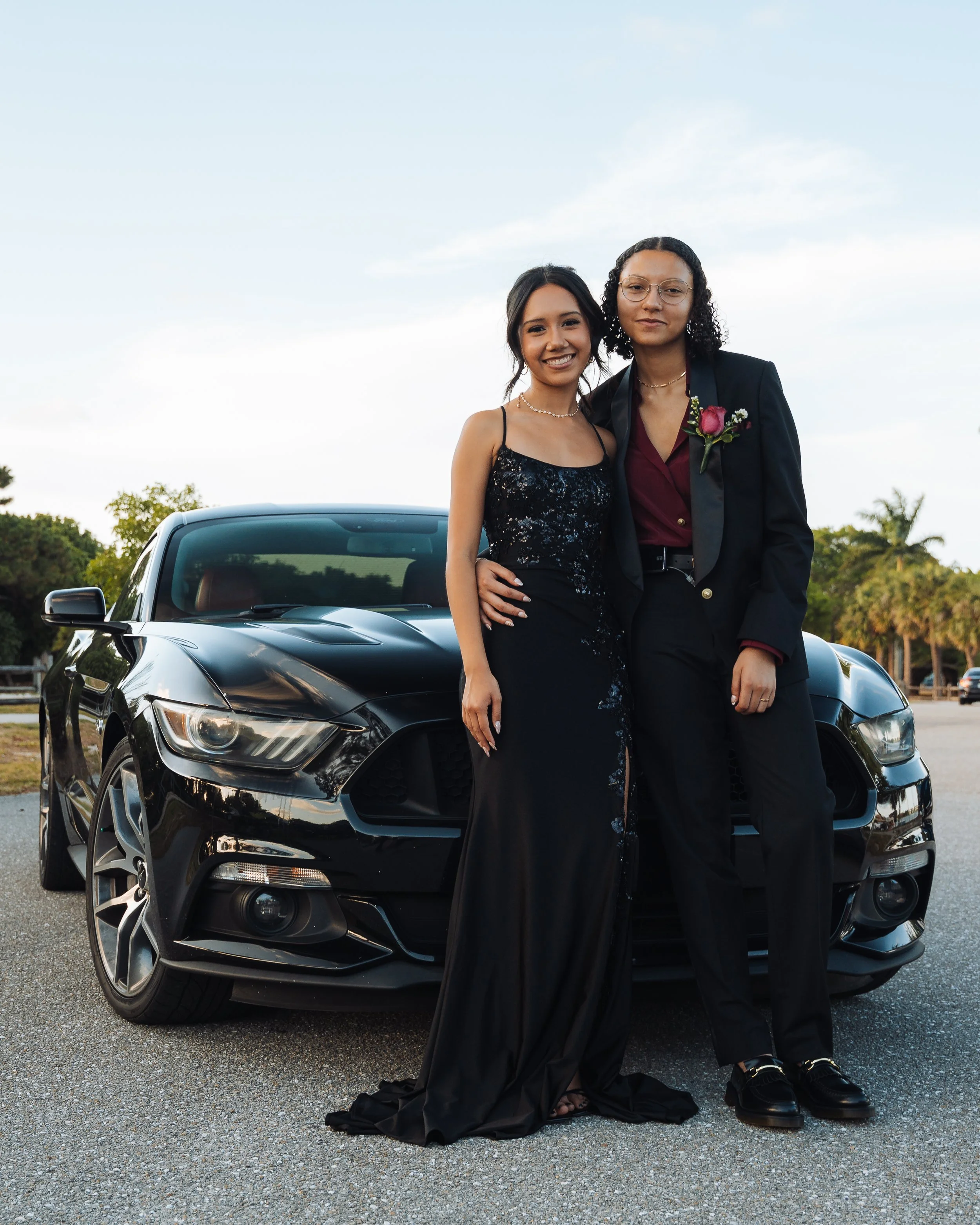 Two women dressed in formal attire standing in front of a black car outdoors, smiling at the camera during sunset.