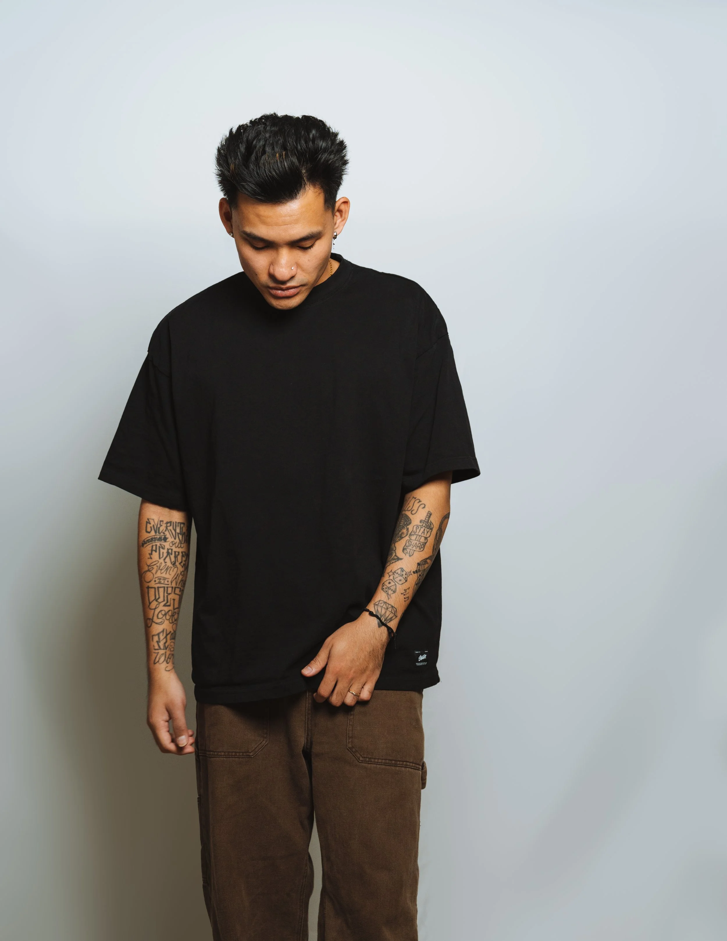 A young man dressed in a black t-shirt and brown pants standing against a plain light-colored wall. He looks down with his left hand resting on his thigh and has tattoos on both arms.
